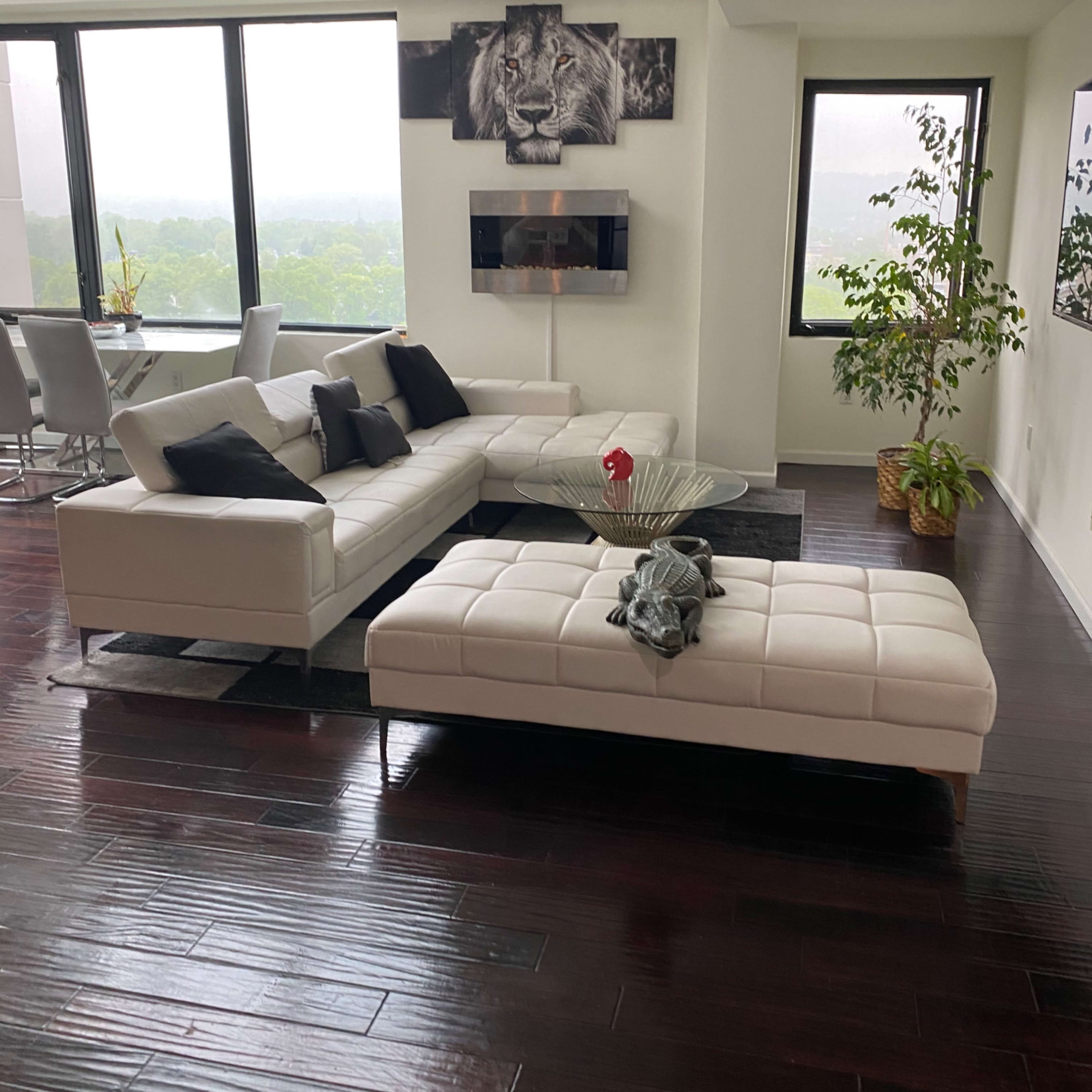 A modern living room with a white sectional sofa, a glass coffee table, and large windows offering a view of greenery outside.