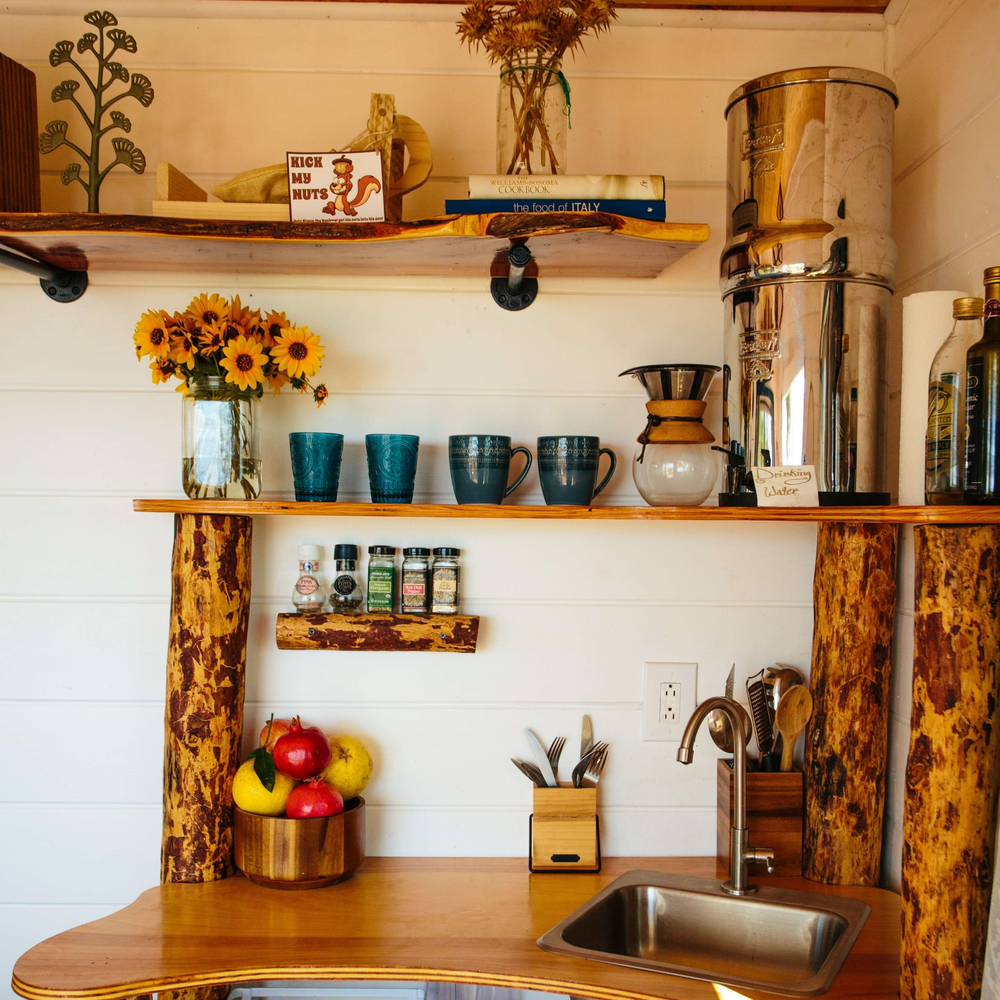A small kitchen area with wooden shelves, a sink, coffee maker, and various kitchen items including mugs, spices, and fresh produce.