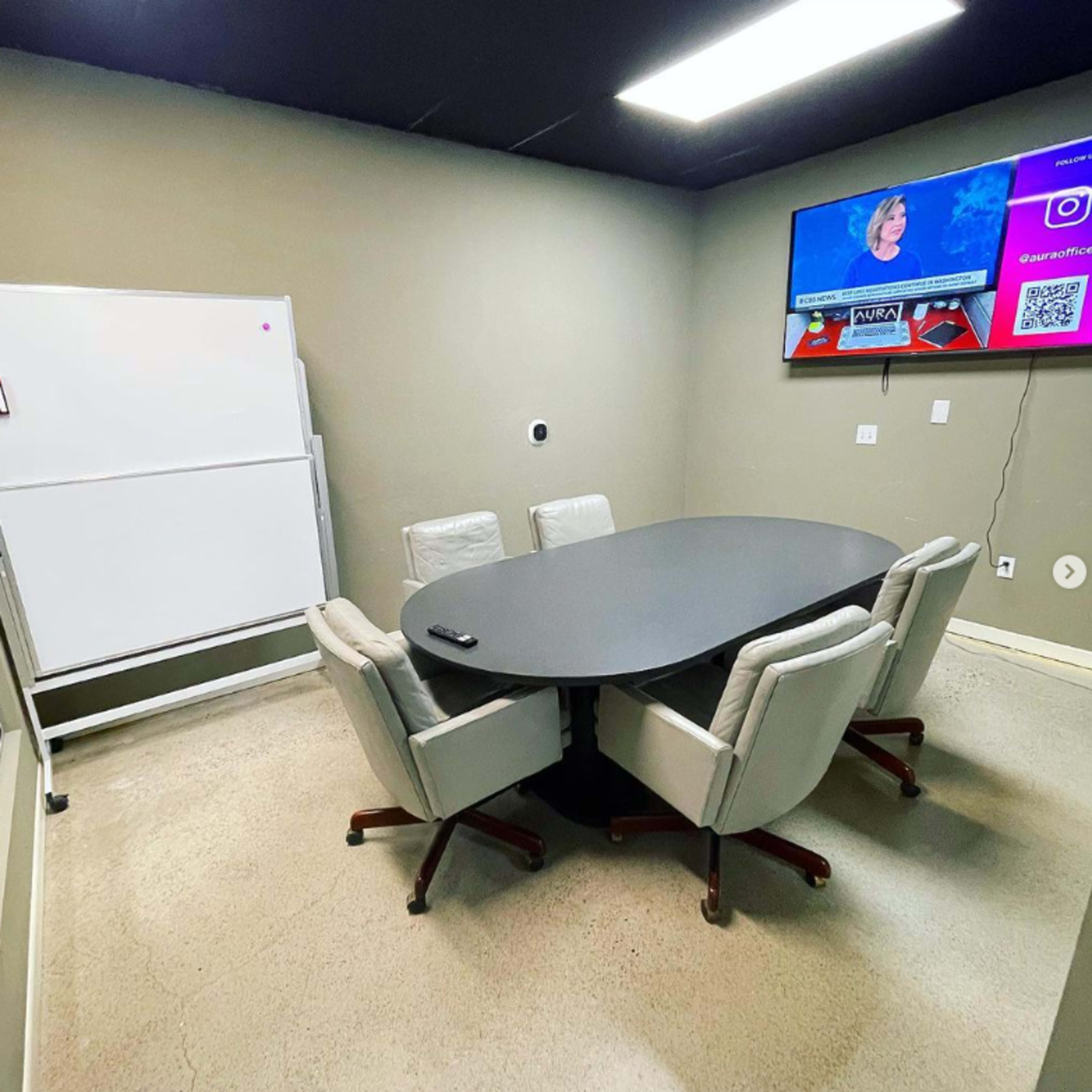 A conference room features a round table surrounded by four chairs, a whiteboard, and a television displaying news content.