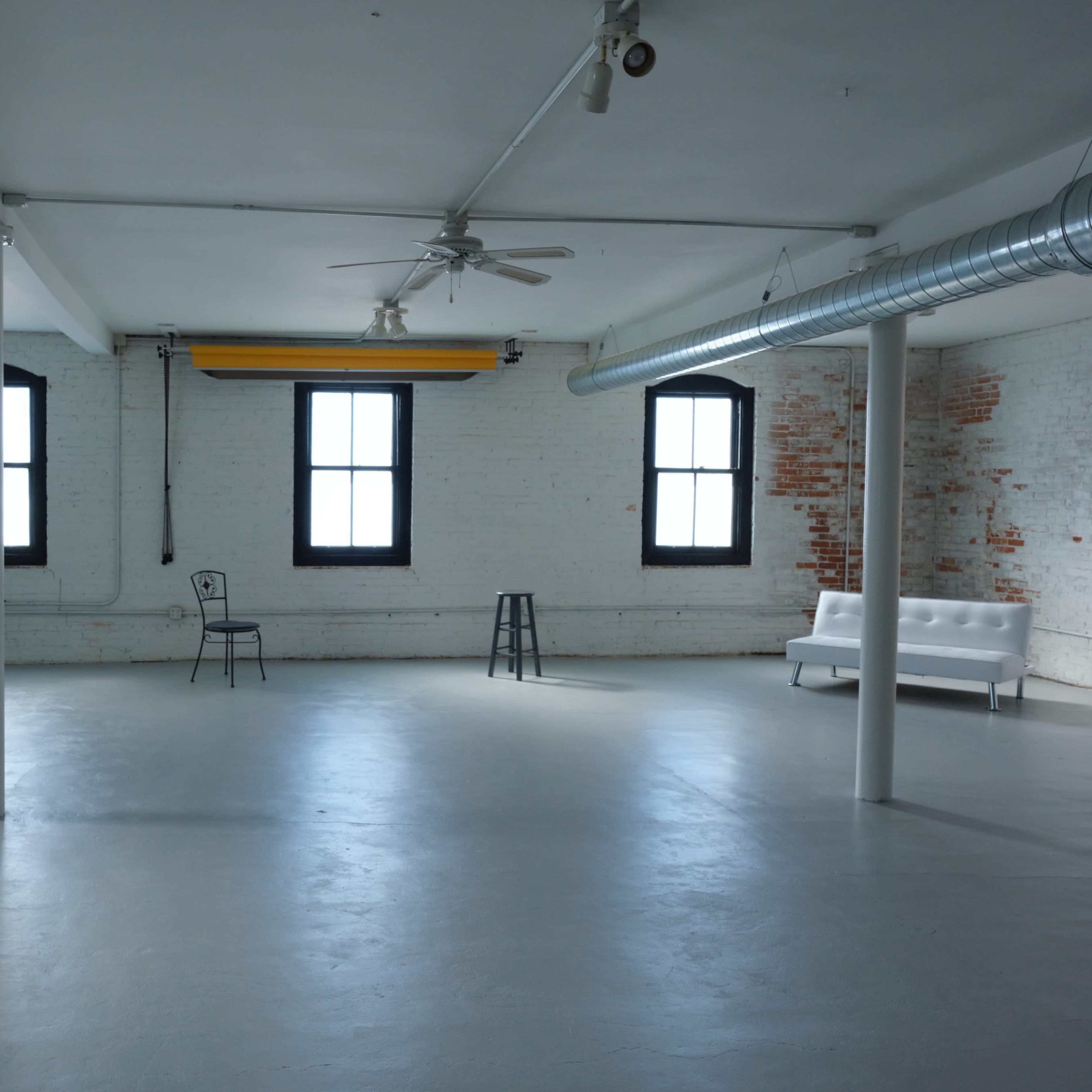 An empty industrial space features white walls, concrete flooring, two windows, a ceiling fan, and a white couch beside two stools.