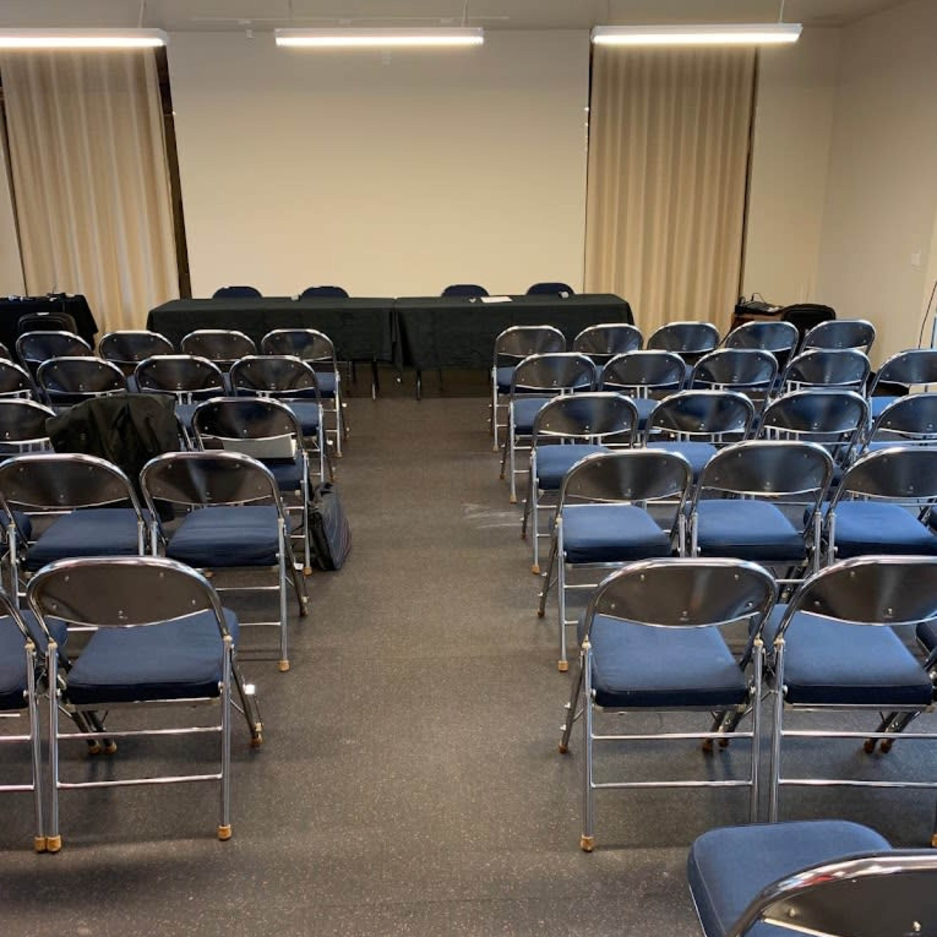 A meeting room with rows of blue chairs facing a table at the front against a light-colored wall.