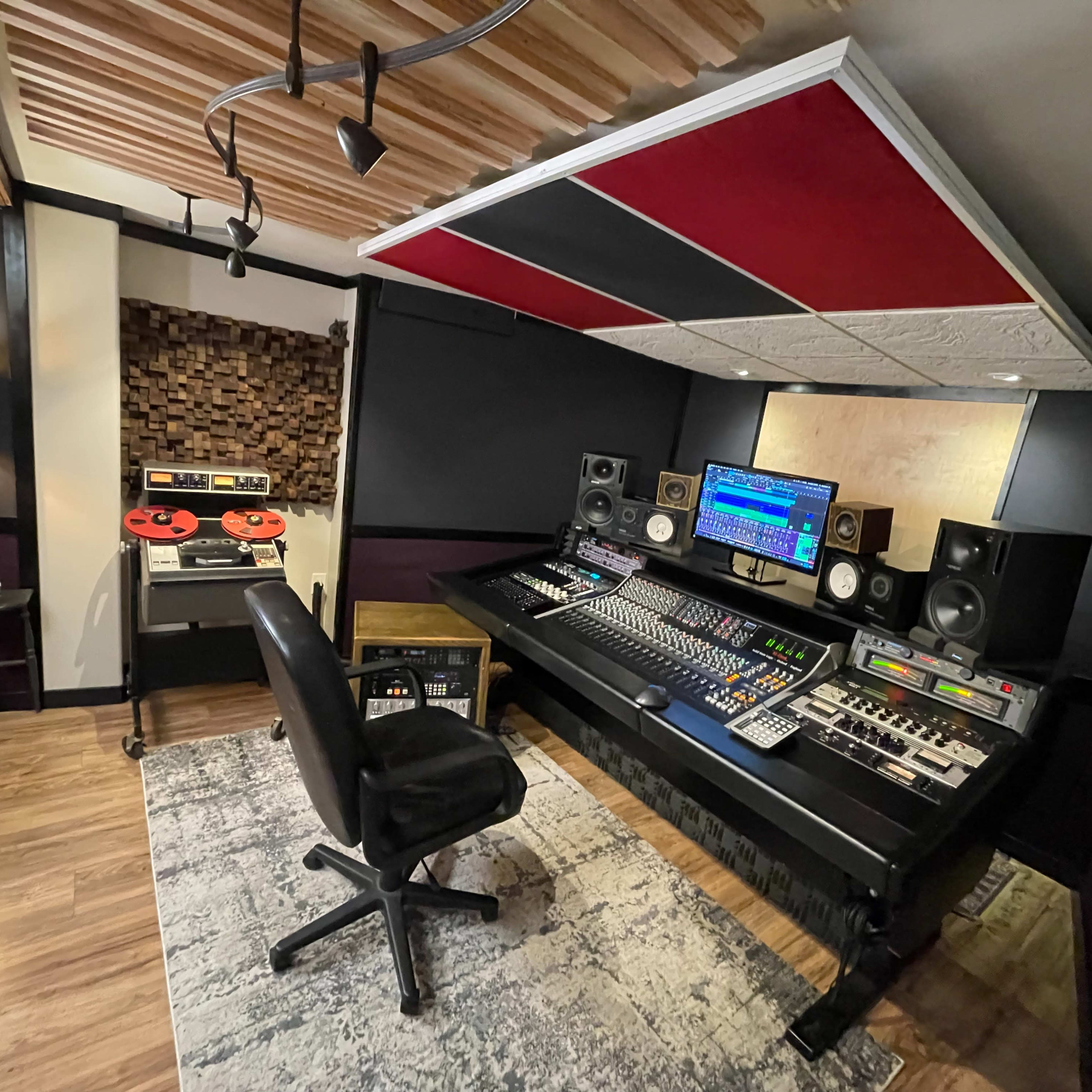 A music studio with a large mixing console, computer monitors, speakers, and soundproof walls, accompanied by a vintage tape deck and an area rug.