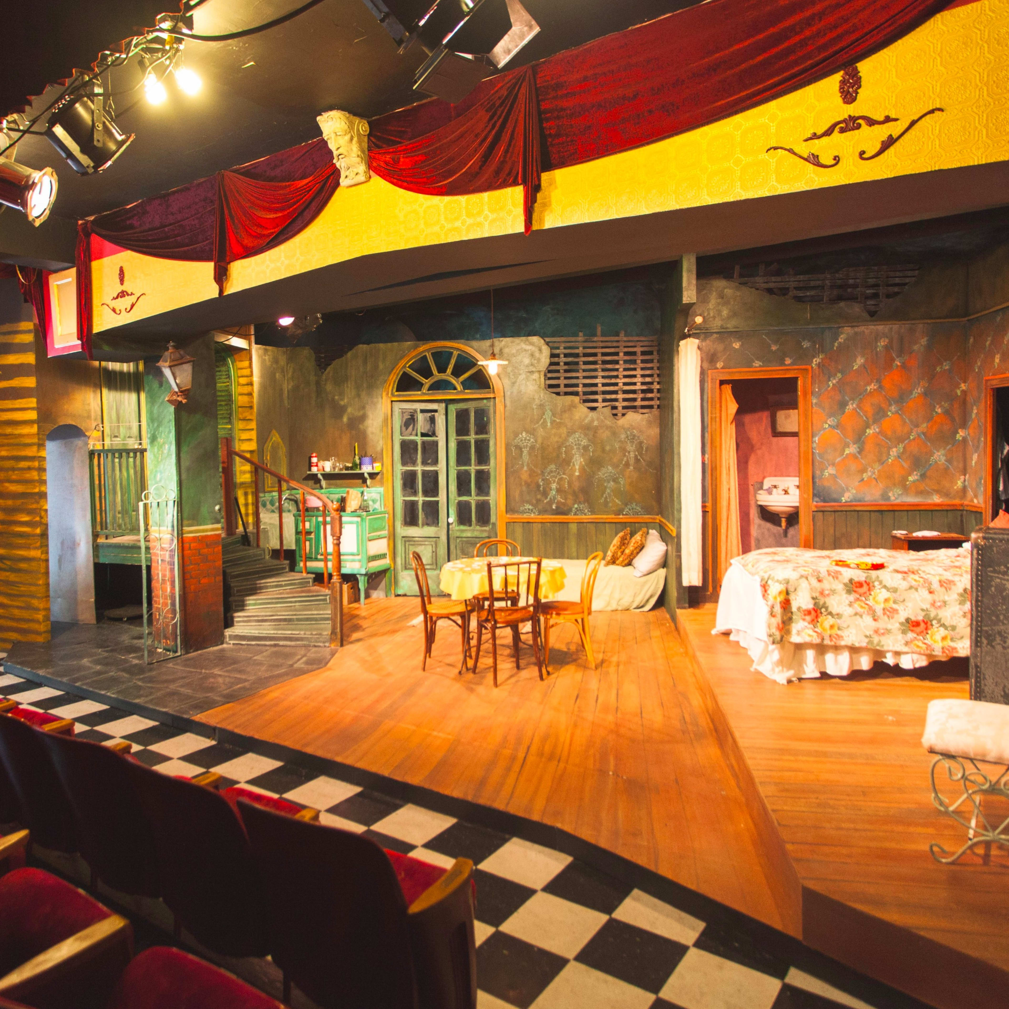 A theater stage set with a colorful backdrop, furniture, and stage lighting, viewed from the audience seating area.