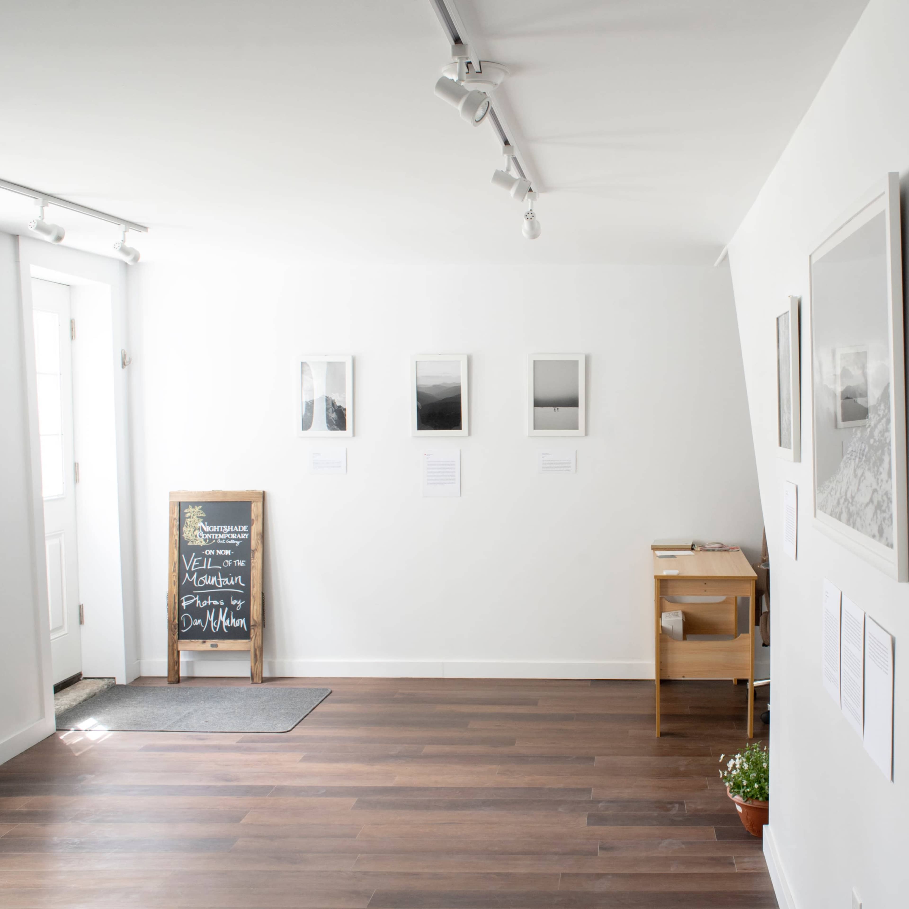 A minimalist gallery space with framed black-and-white photographs on the wall, a wooden table, and a chalkboard near the entrance.