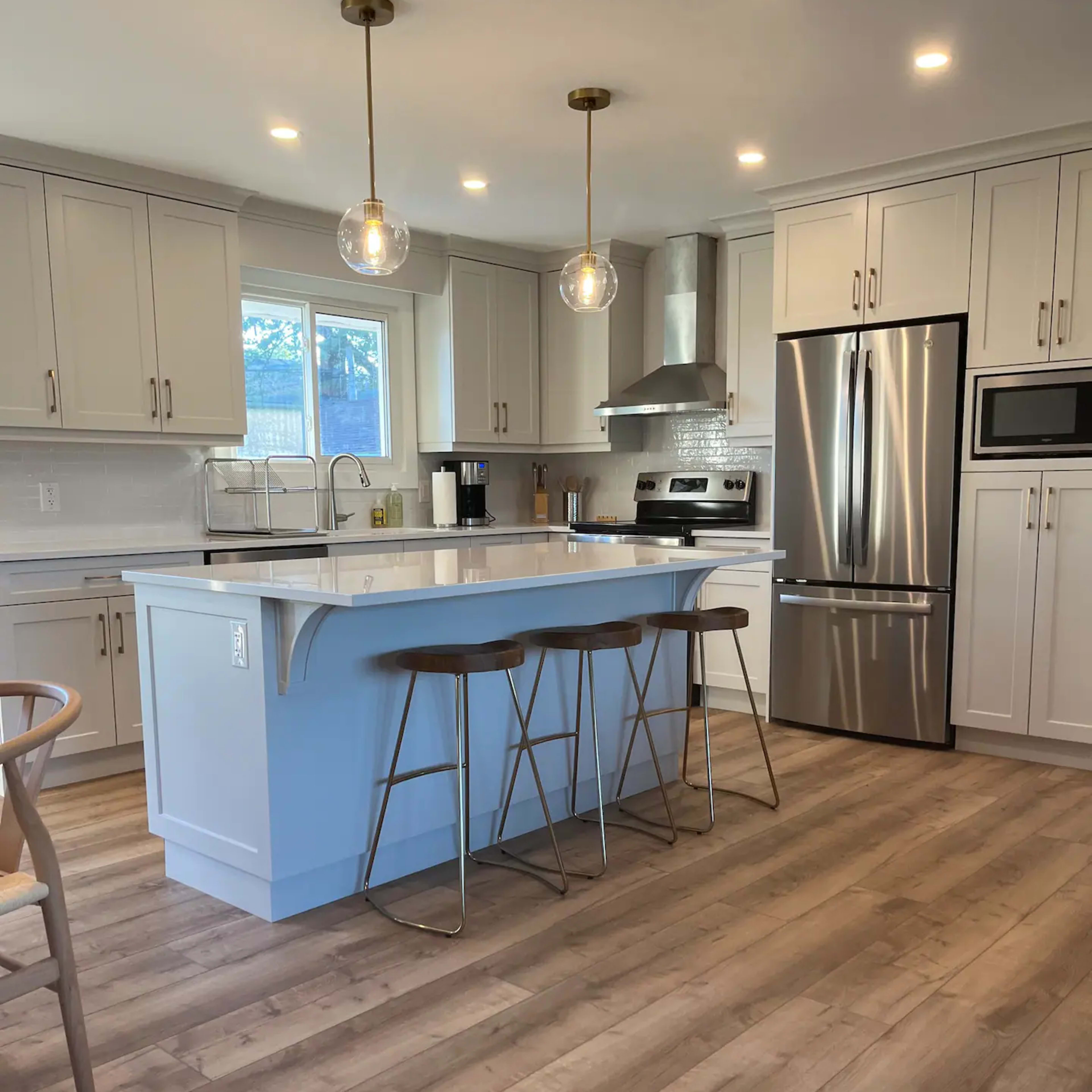 A modern kitchen features light cabinetry, a large island with three bar stools, and stainless steel appliances.