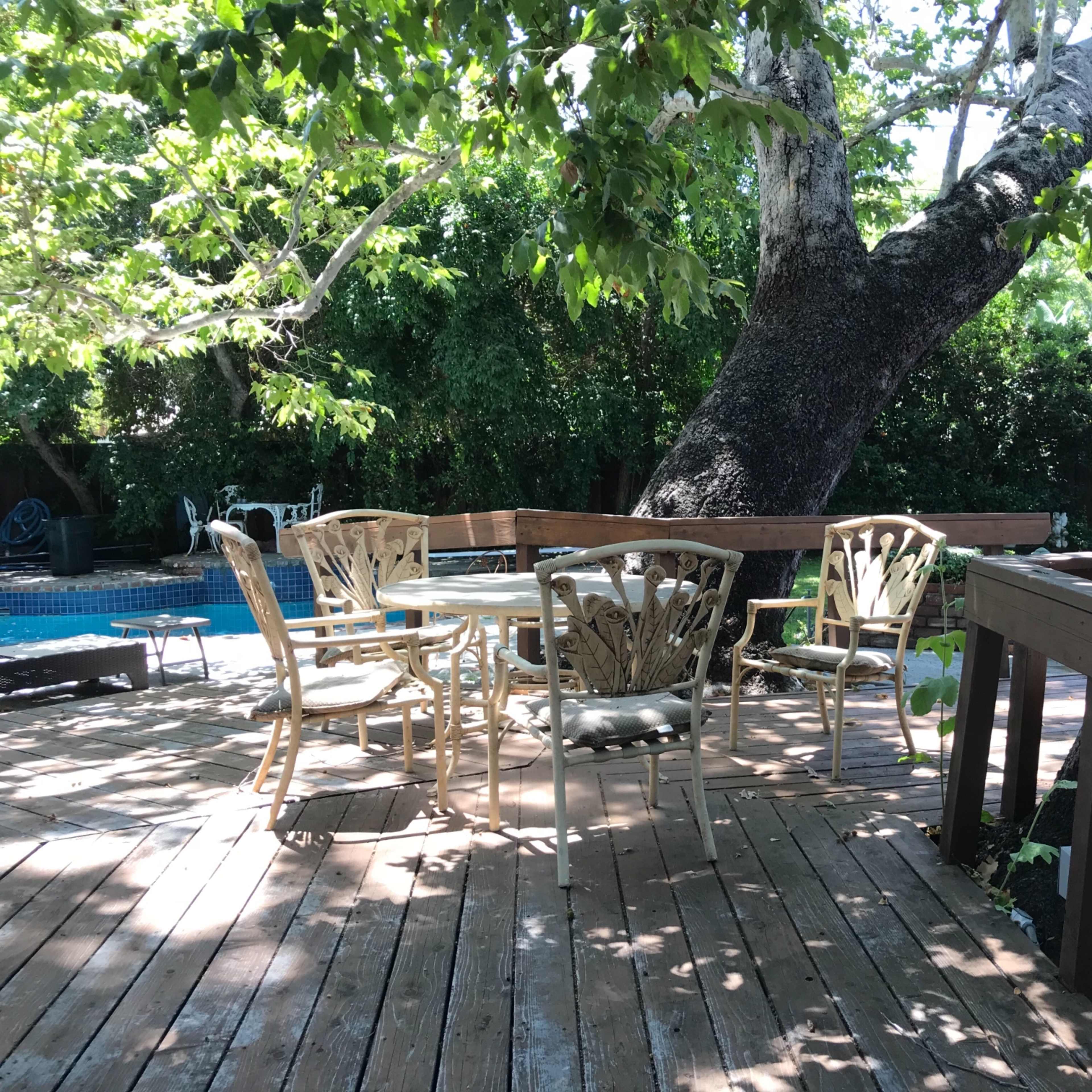 A wooden deck features white outdoor furniture surrounded by trees, with a swimming pool visible in the background.