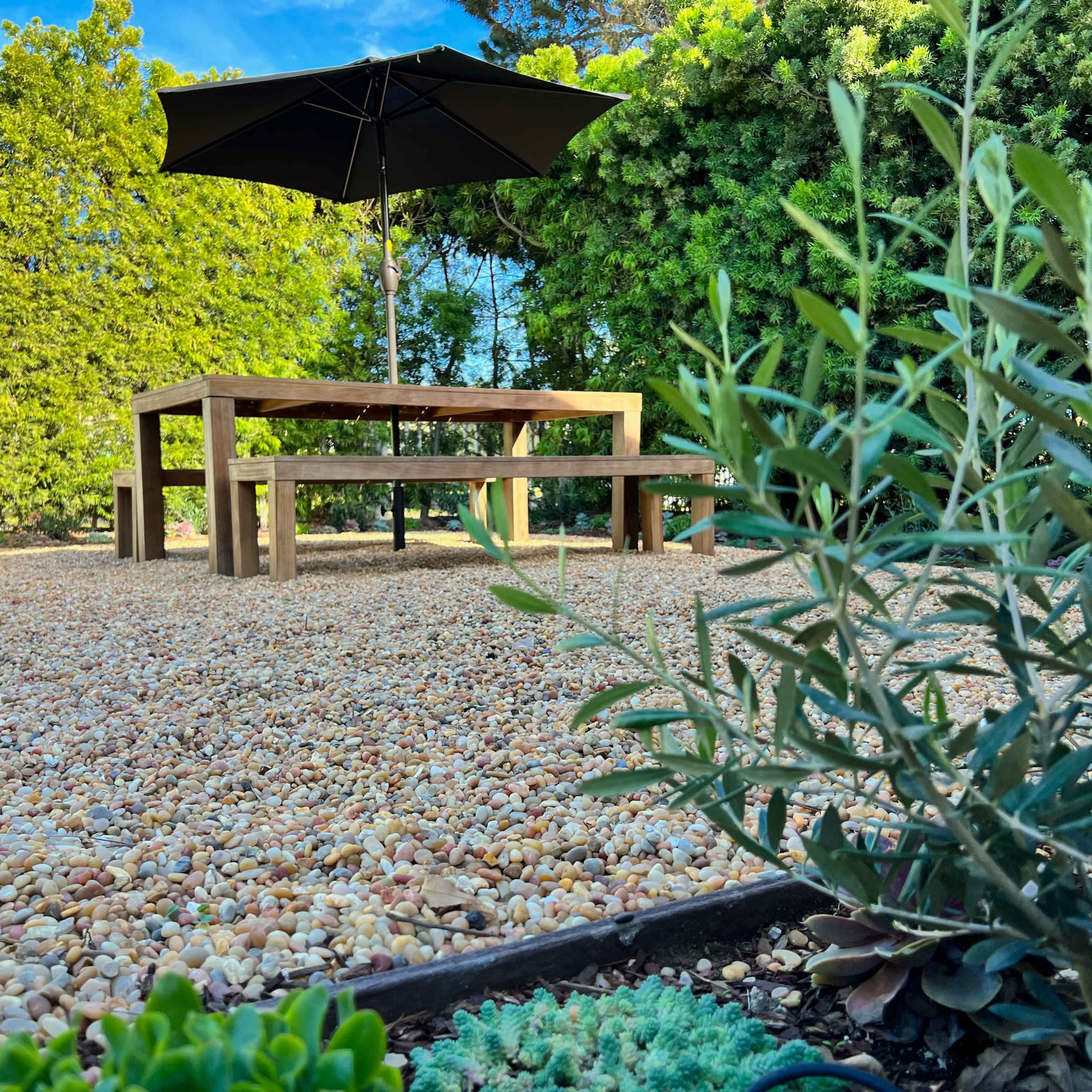 A wooden table and bench set sits on a gravel surface under a large black umbrella, surrounded by greenery.