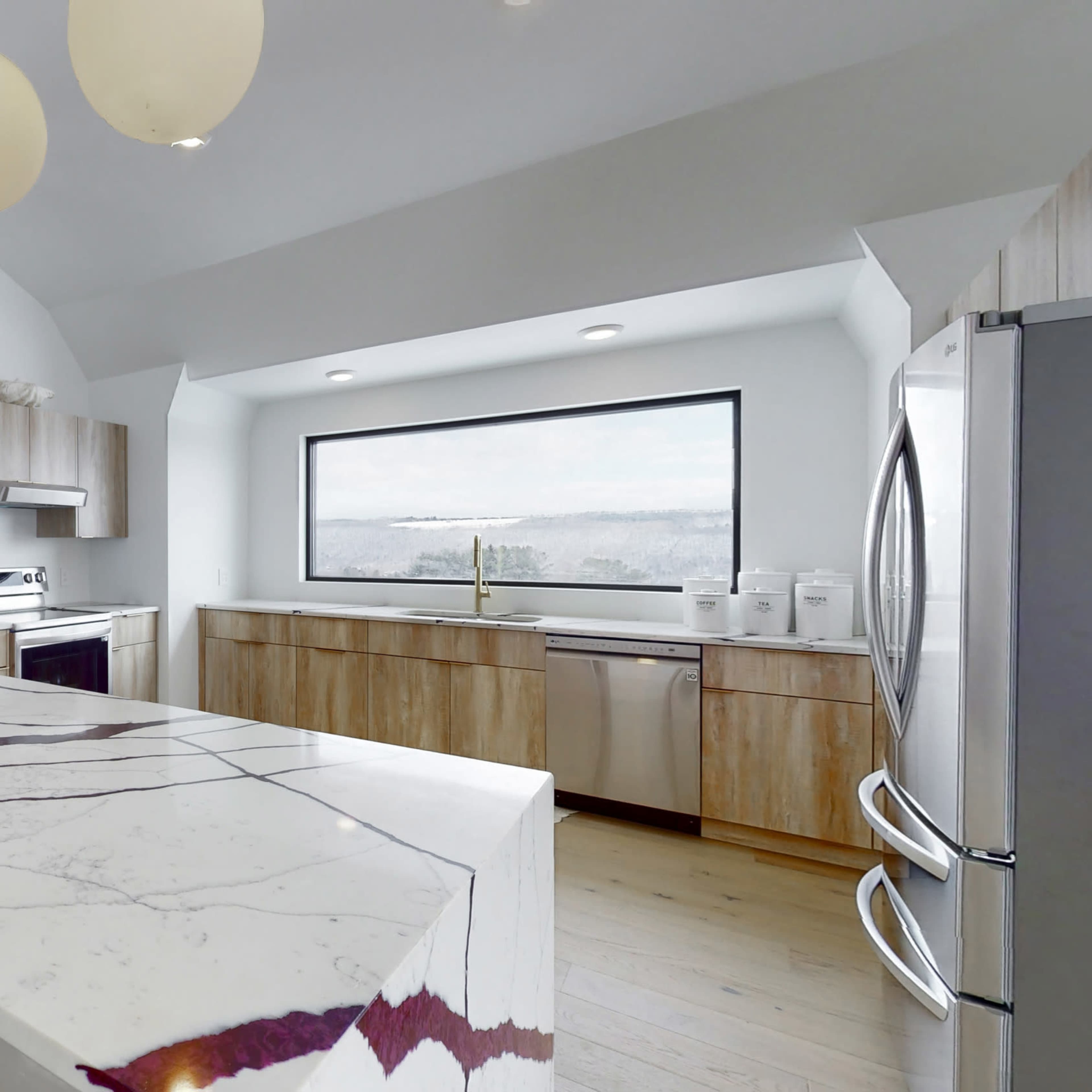 A modern kitchen features a large island with a white and purple marble countertop, wooden cabinetry, and large windows offering a scenic view.