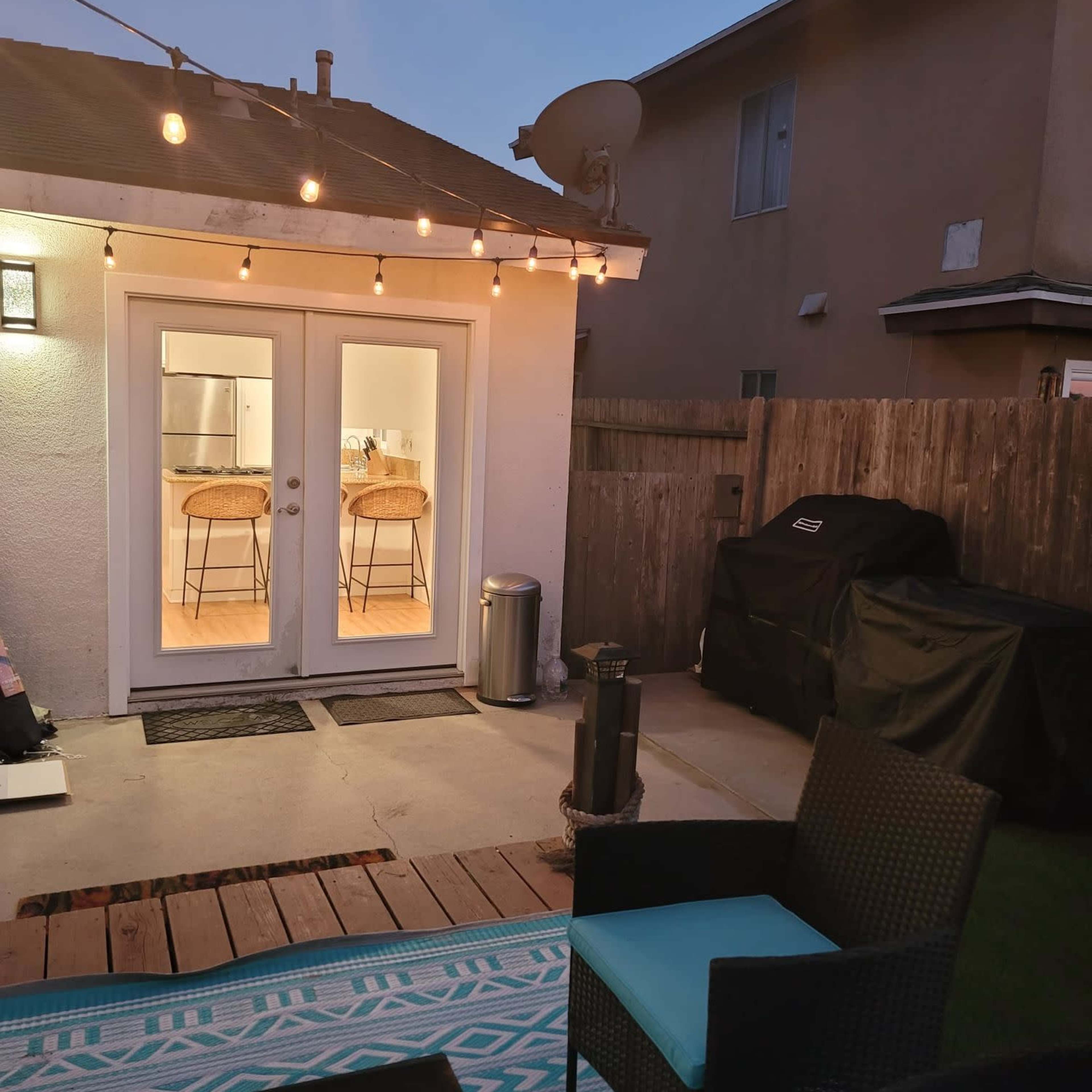 A backyard patio with string lights, a pair of chairs, a small table, and a view of a kitchen through double glass doors.