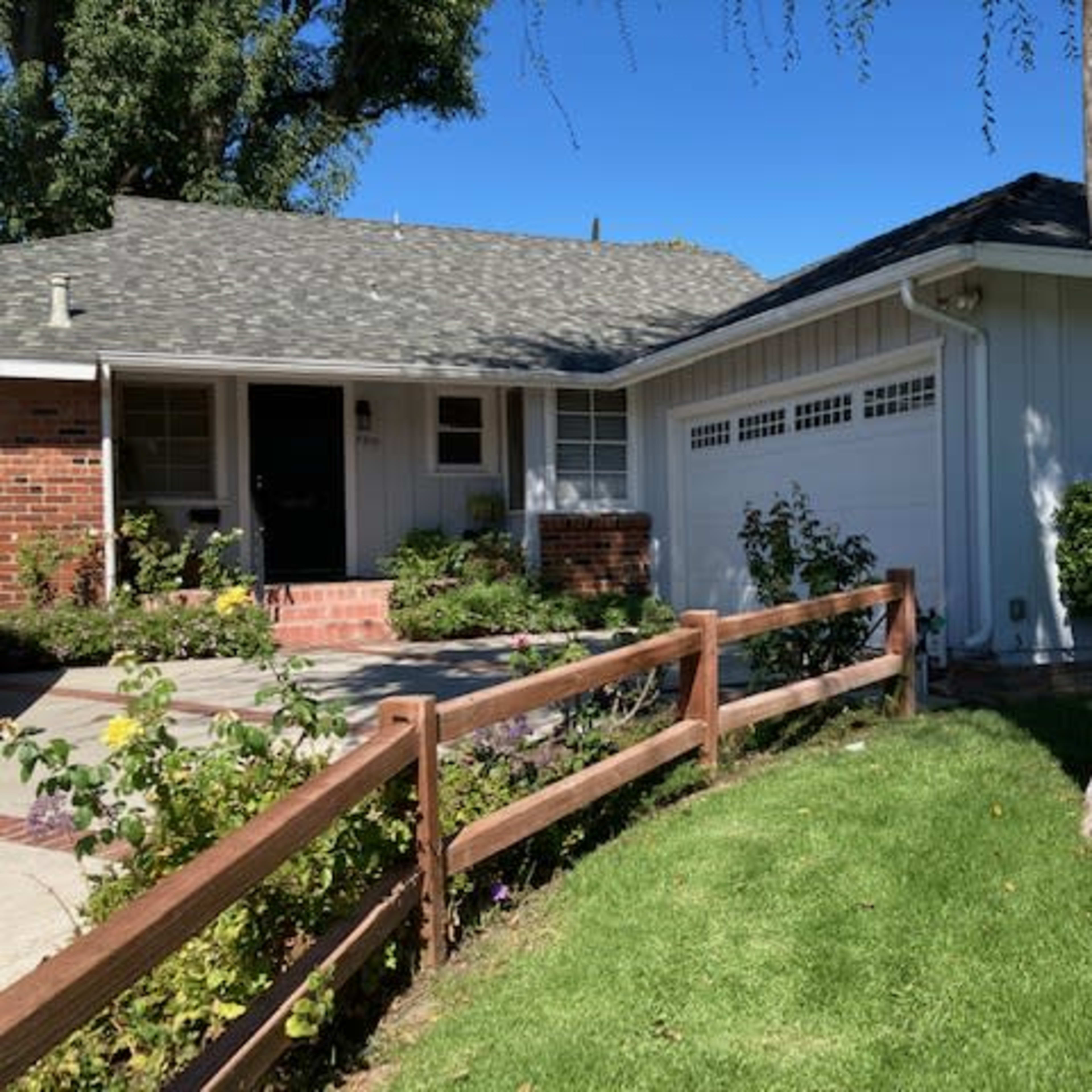 A single-story house with a shingled roof and a white exterior is surrounded by a manicured lawn and a wooden fence.
