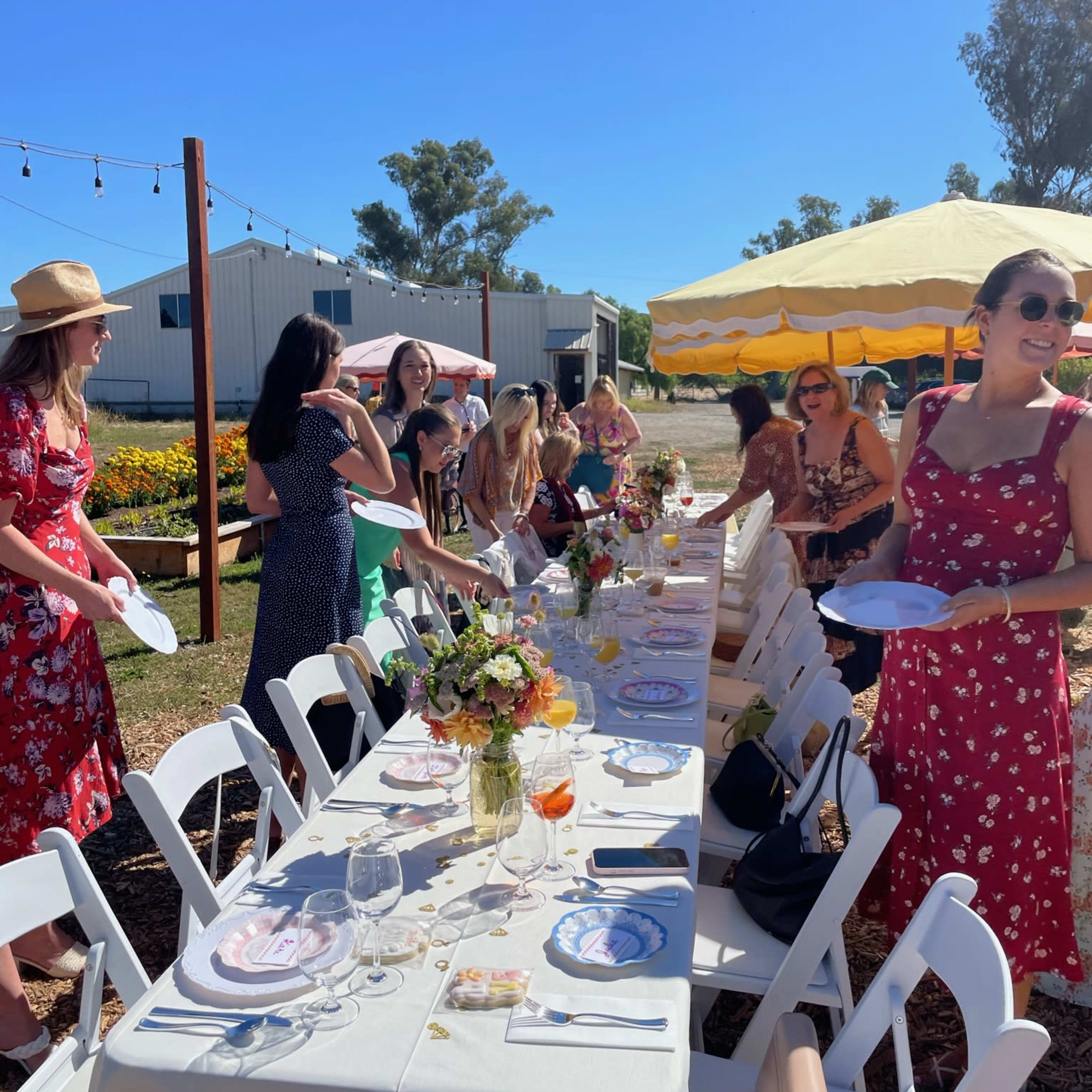 A long outdoor dining table is set for a gathering, surrounded by women dressed in colorful attire, with a tent and flowers in the background under a clear blue sky.