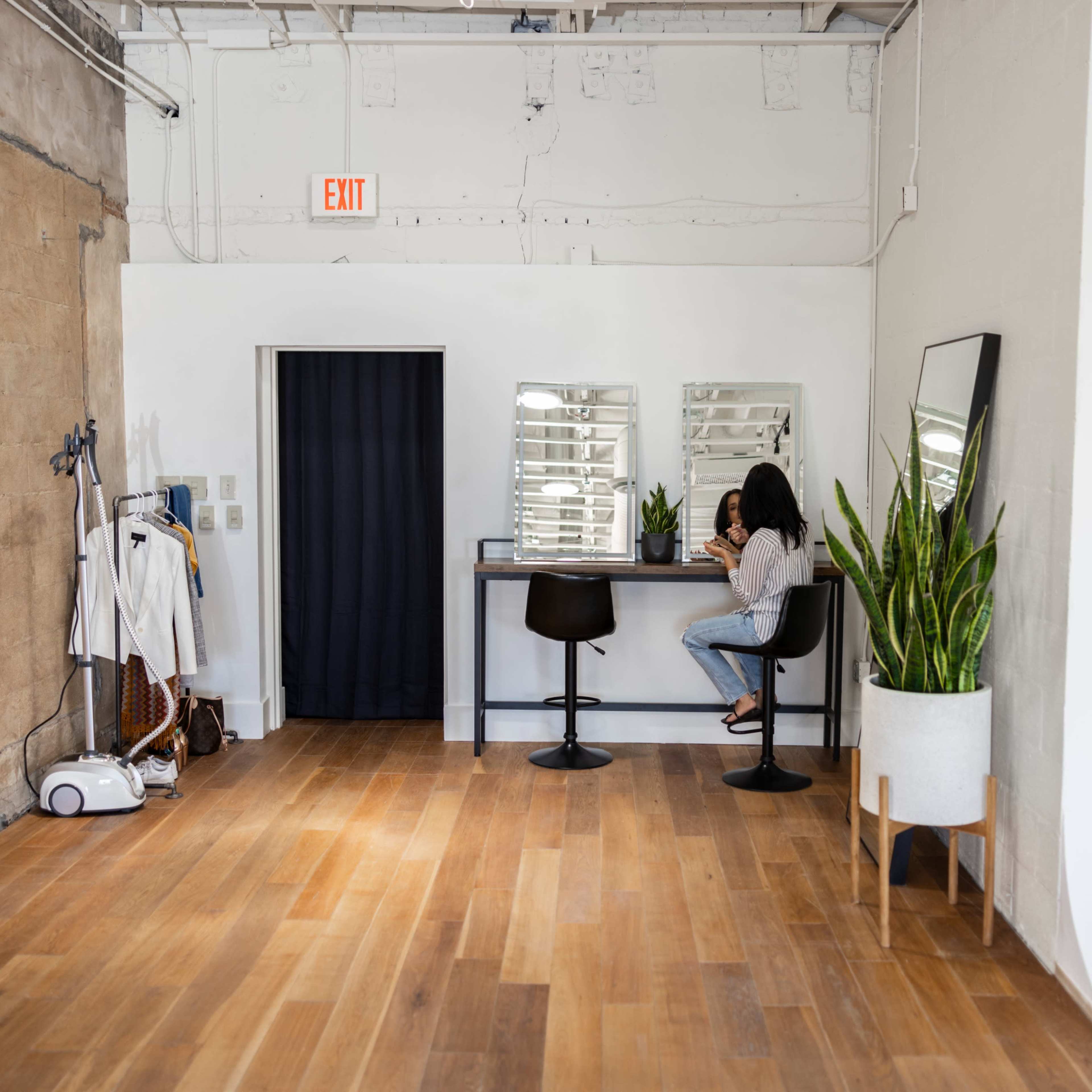 A person sits at a bar-height table in a minimalist room with large mirrors and potted plants, while a small vacuum and clothing rack are visible against the wall.