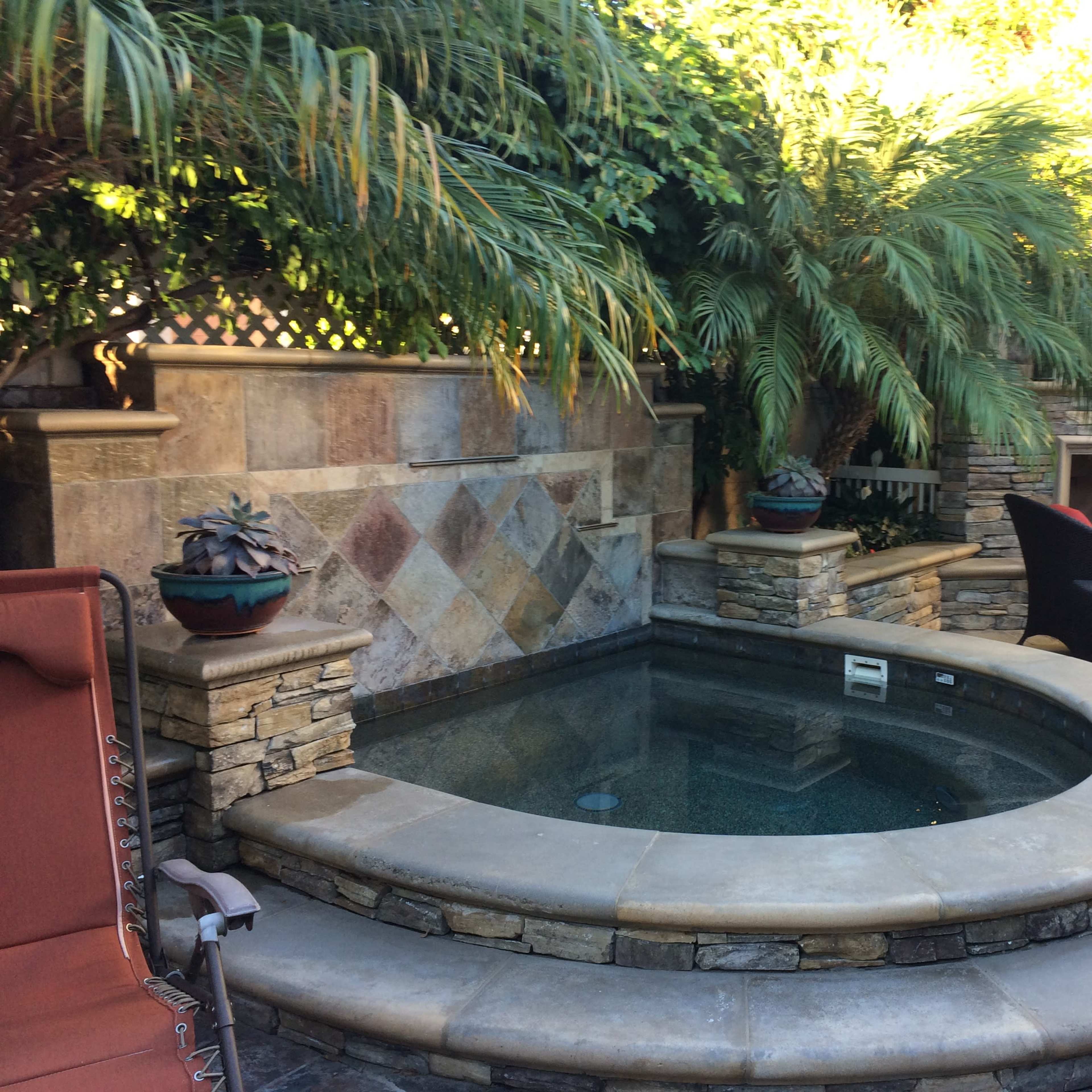 A circular hot tub surrounded by stone walls and tropical plants, with two black chairs and a terracotta-colored lounger nearby.