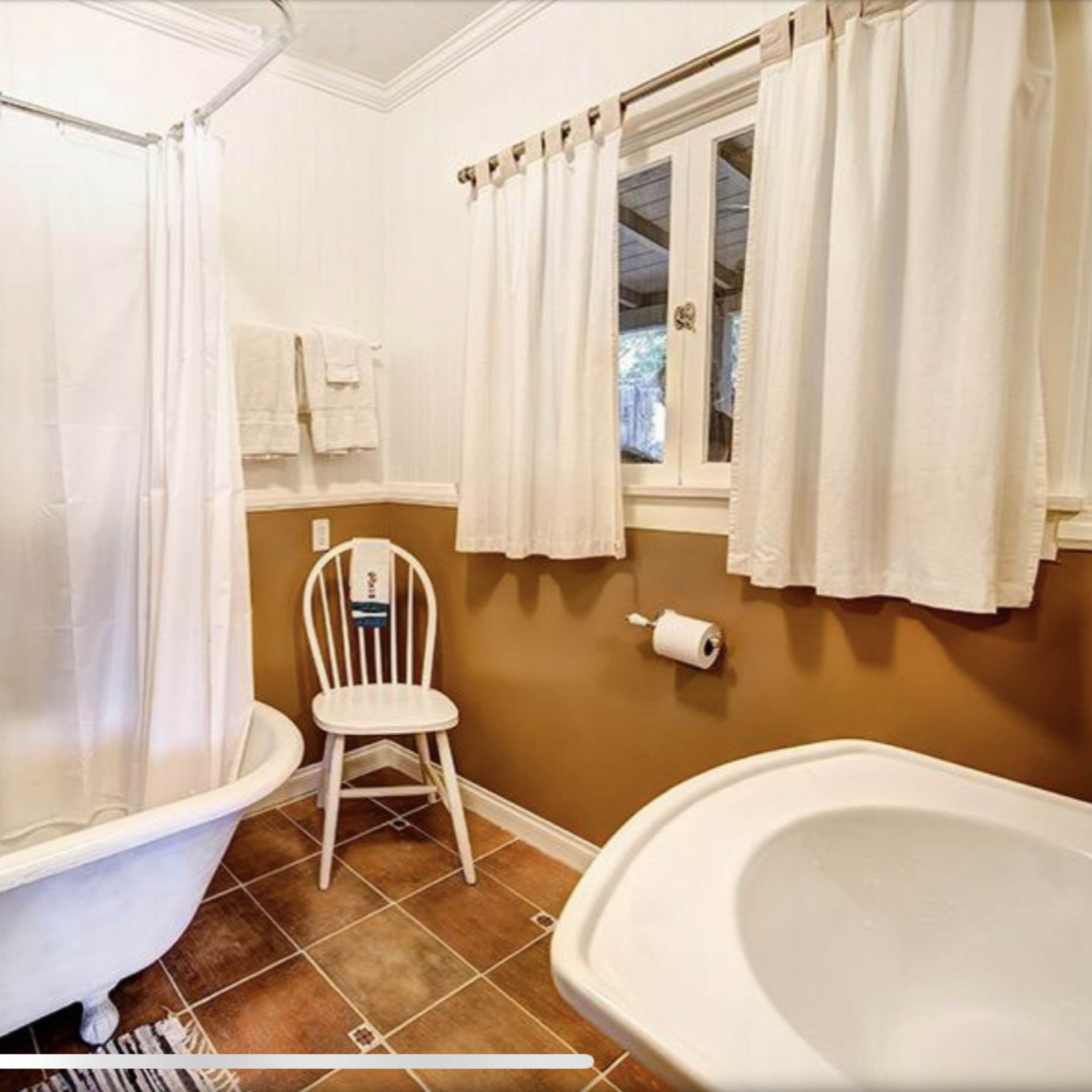 A bathroom with a vintage bathtub, a white shower curtain, a chair, and a window with light-colored curtains.