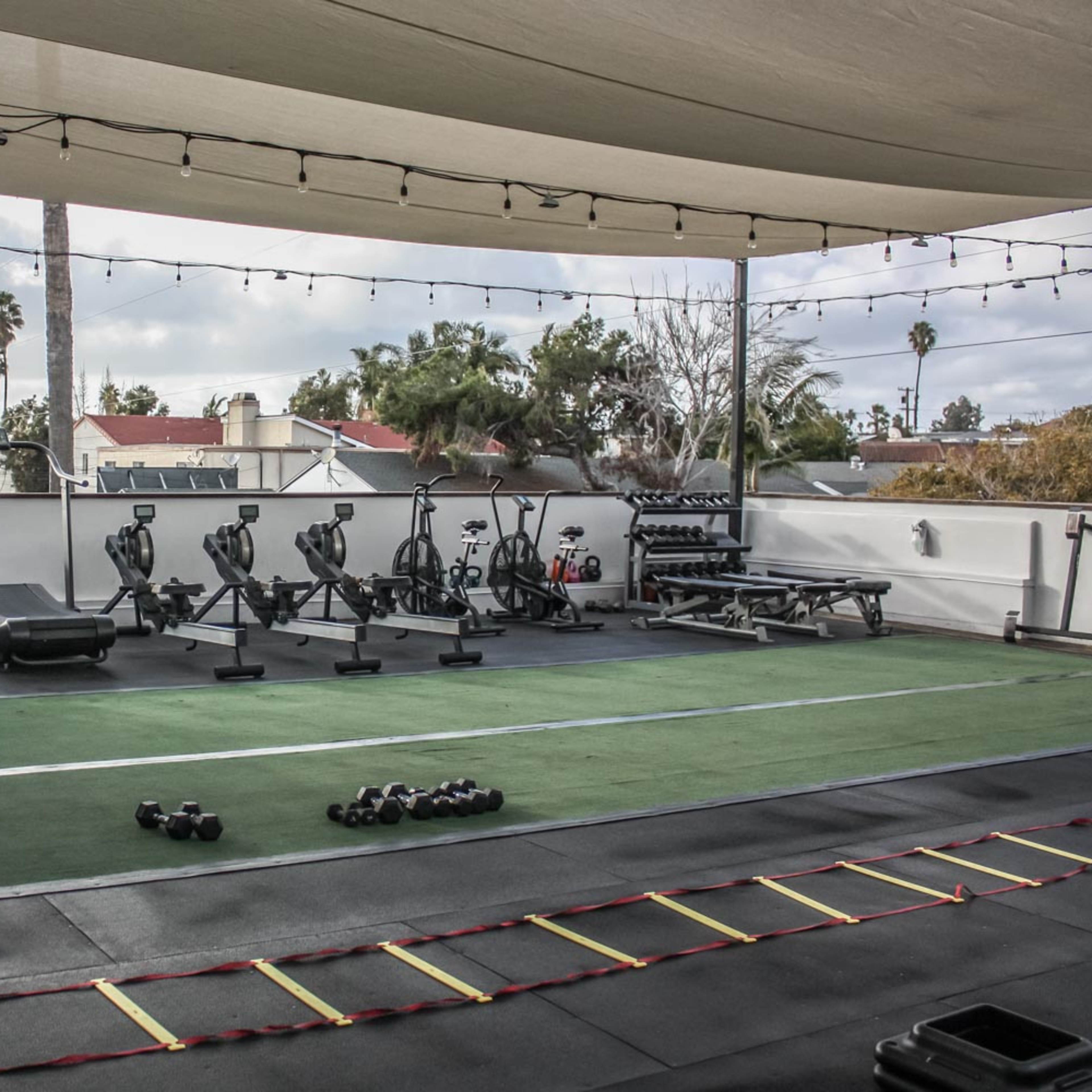 A rooftop gym equipped with various fitness machines, free weights, and exercise mats under a shaded canopy.