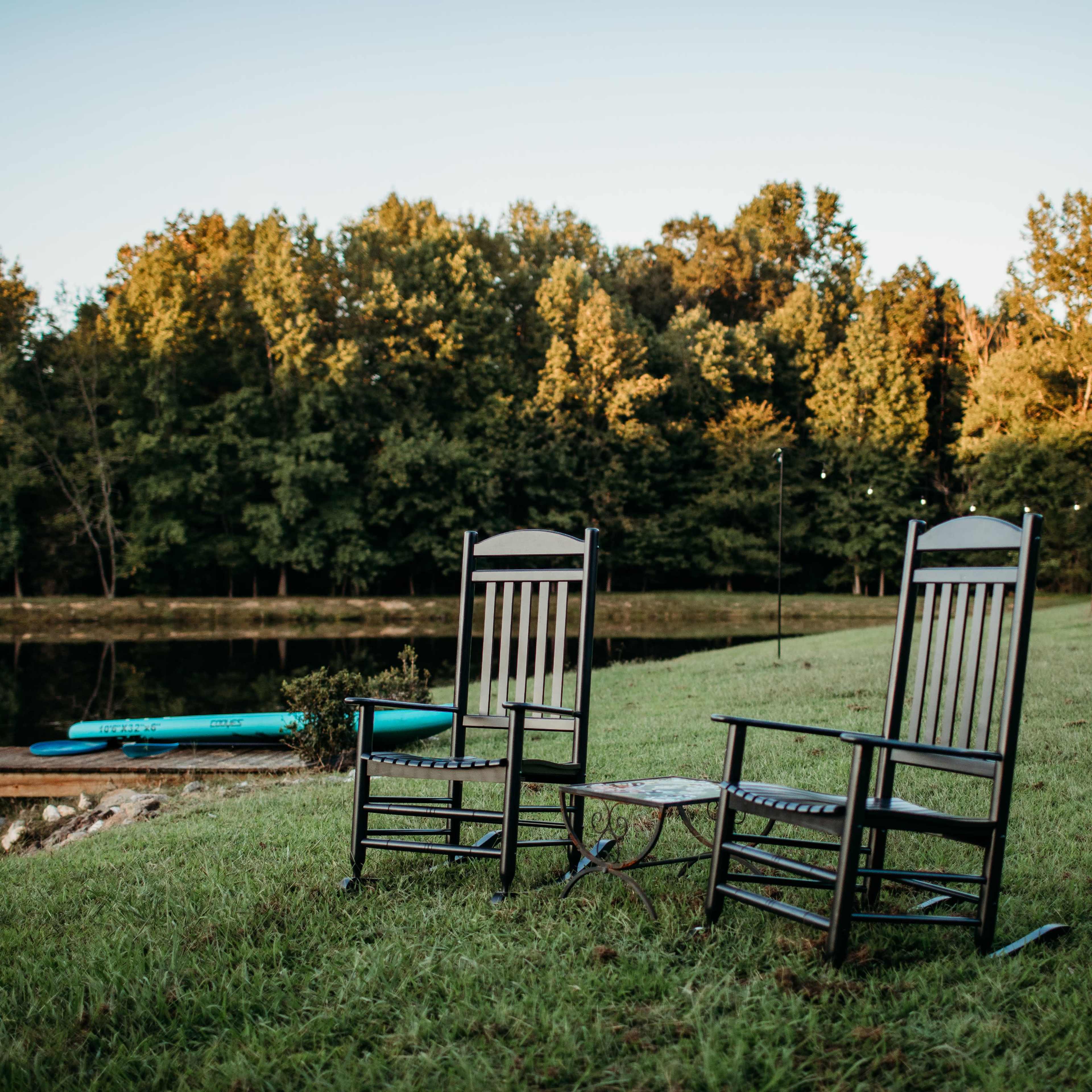 Two rocking chairs and a small table sit on the grass near a calm lake, surrounded by trees.
