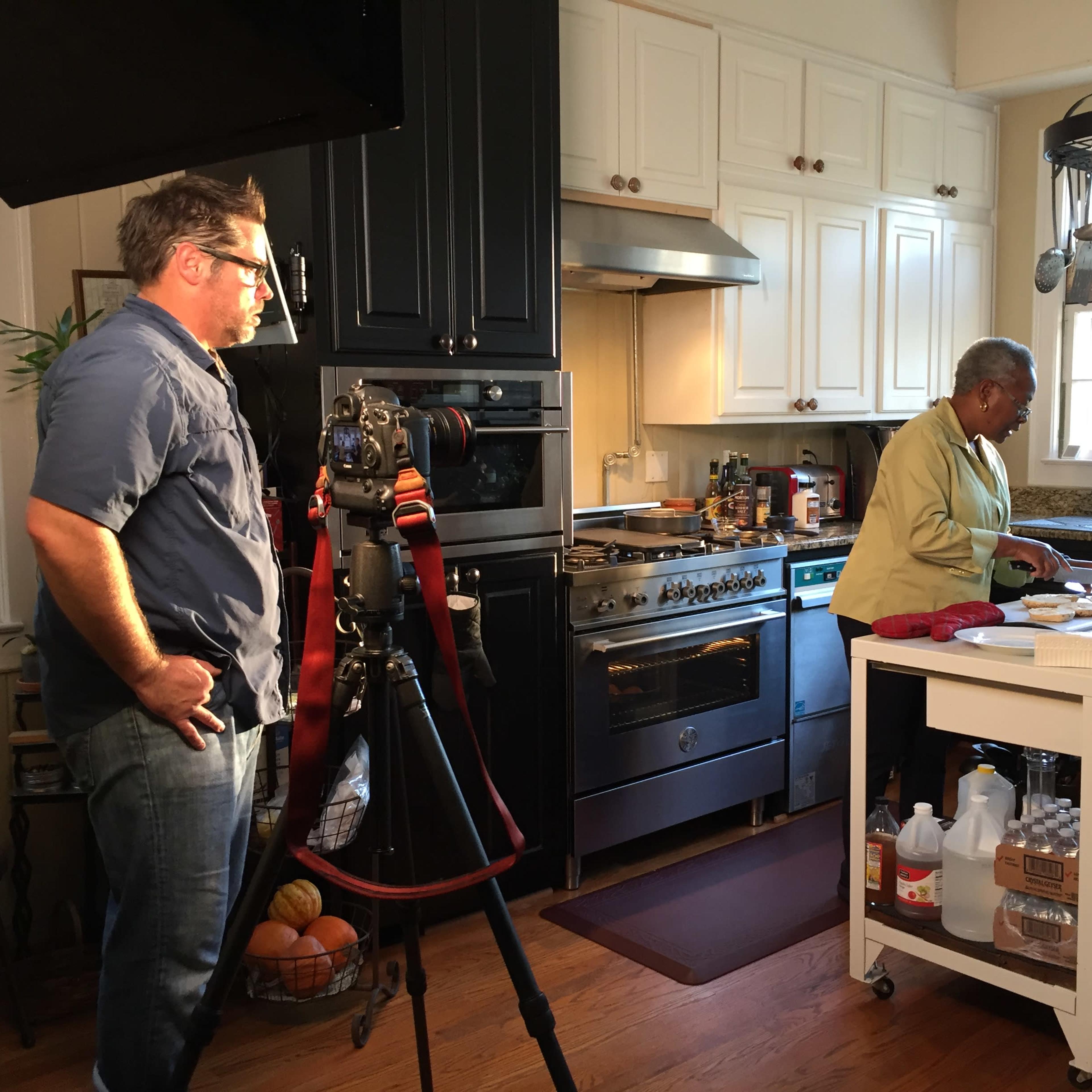 A camera crew sets up to film a cooking segment in a bright kitchen featuring a stovetop and a countertop with various ingredients.