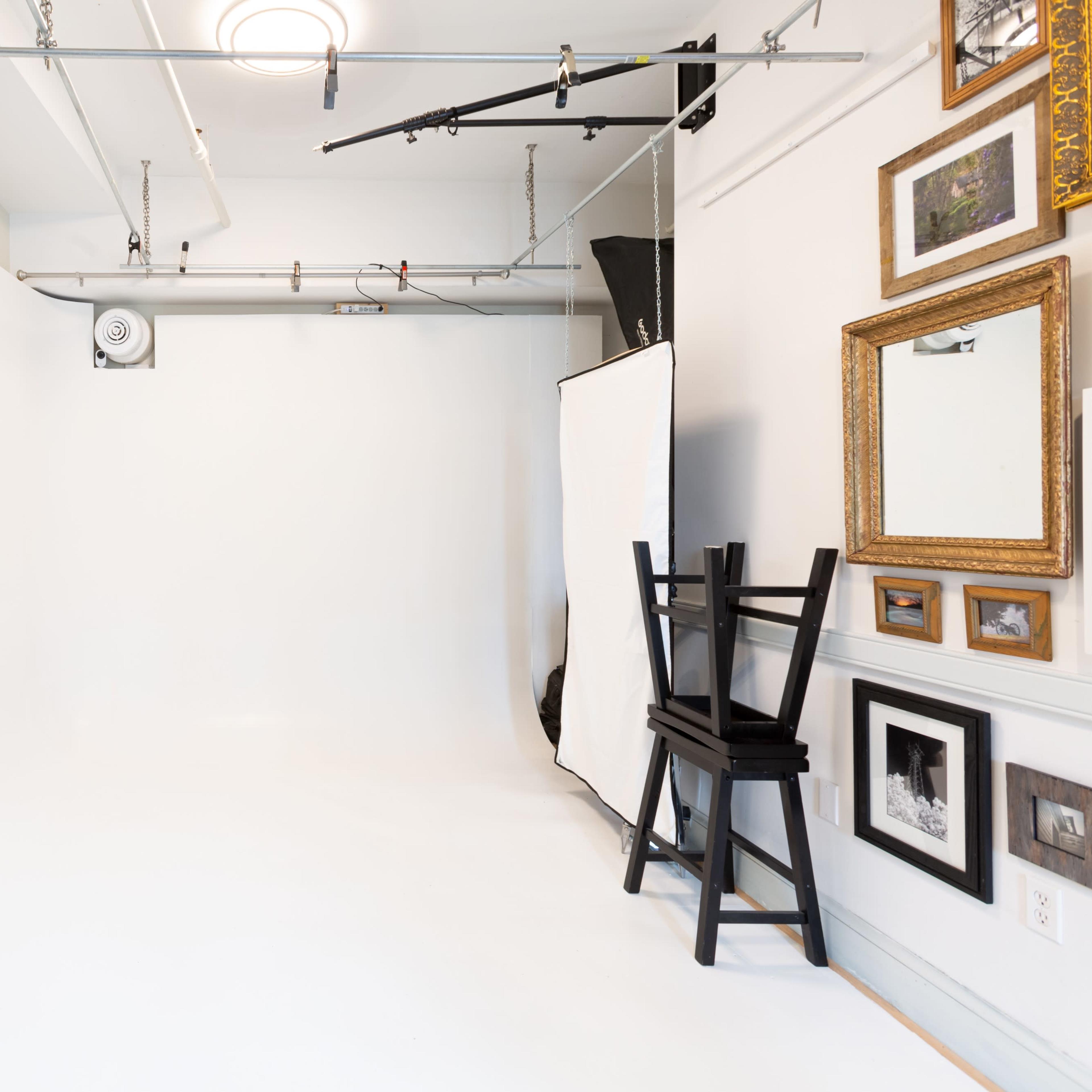 A photography studio features a blank white backdrop, a black chair, and various framed pictures hung on the wall.