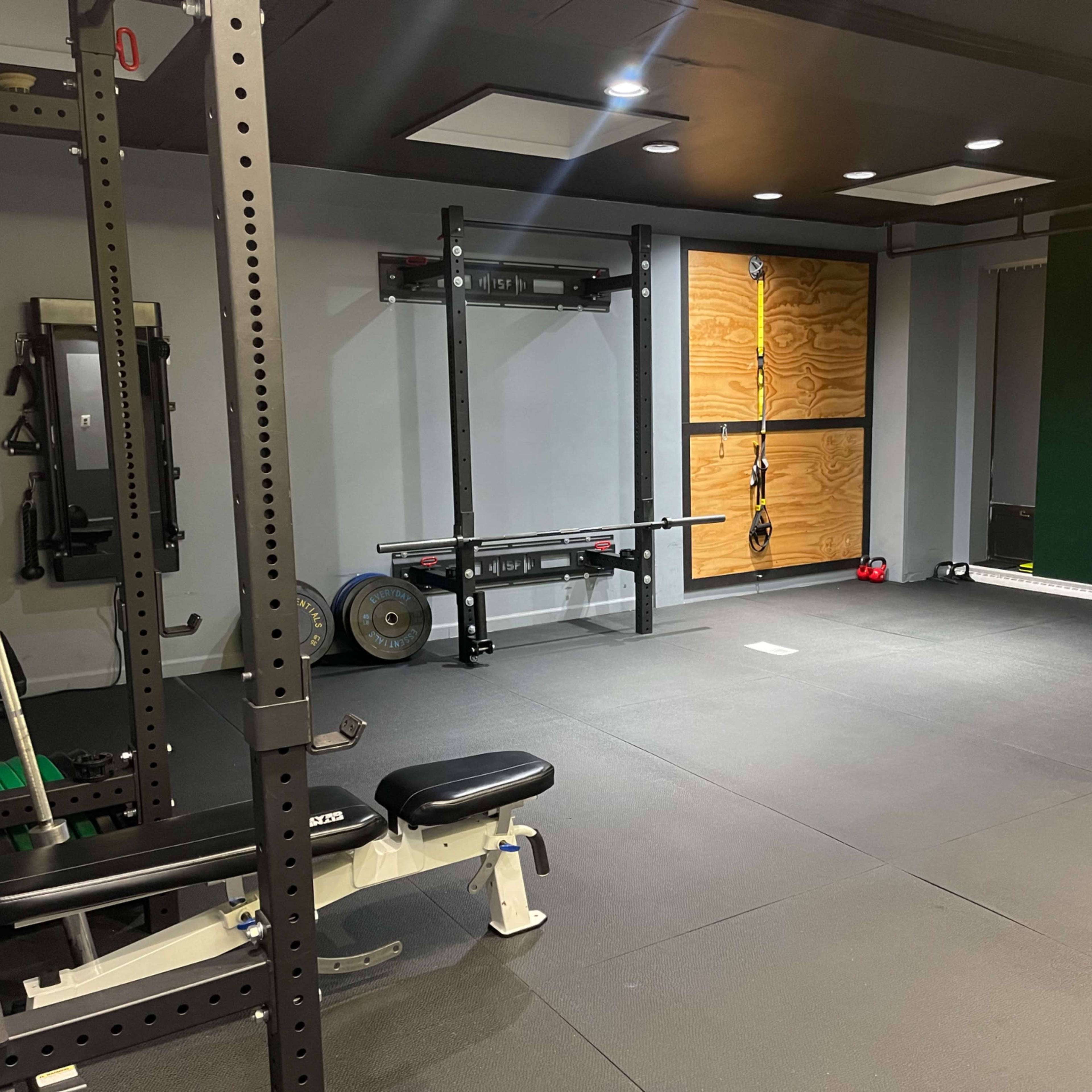 A gym interior featuring a weight bench, squat rack, and wall-mounted exercise equipment in a minimalist space with rubber flooring.