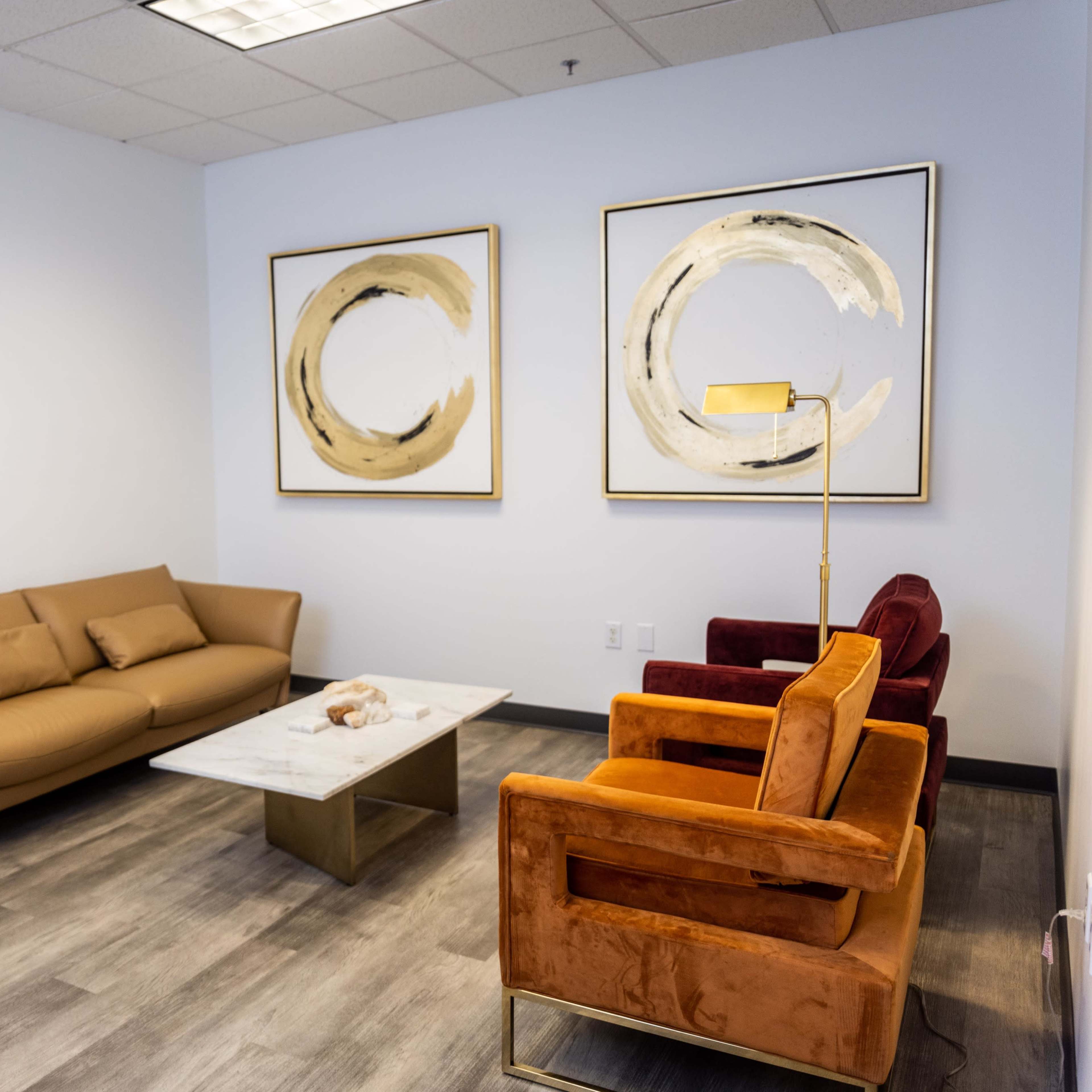 A modern waiting room with a brown sofa, orange and red armchairs, a white marble coffee table, and two abstract gold-framed artworks on the wall.