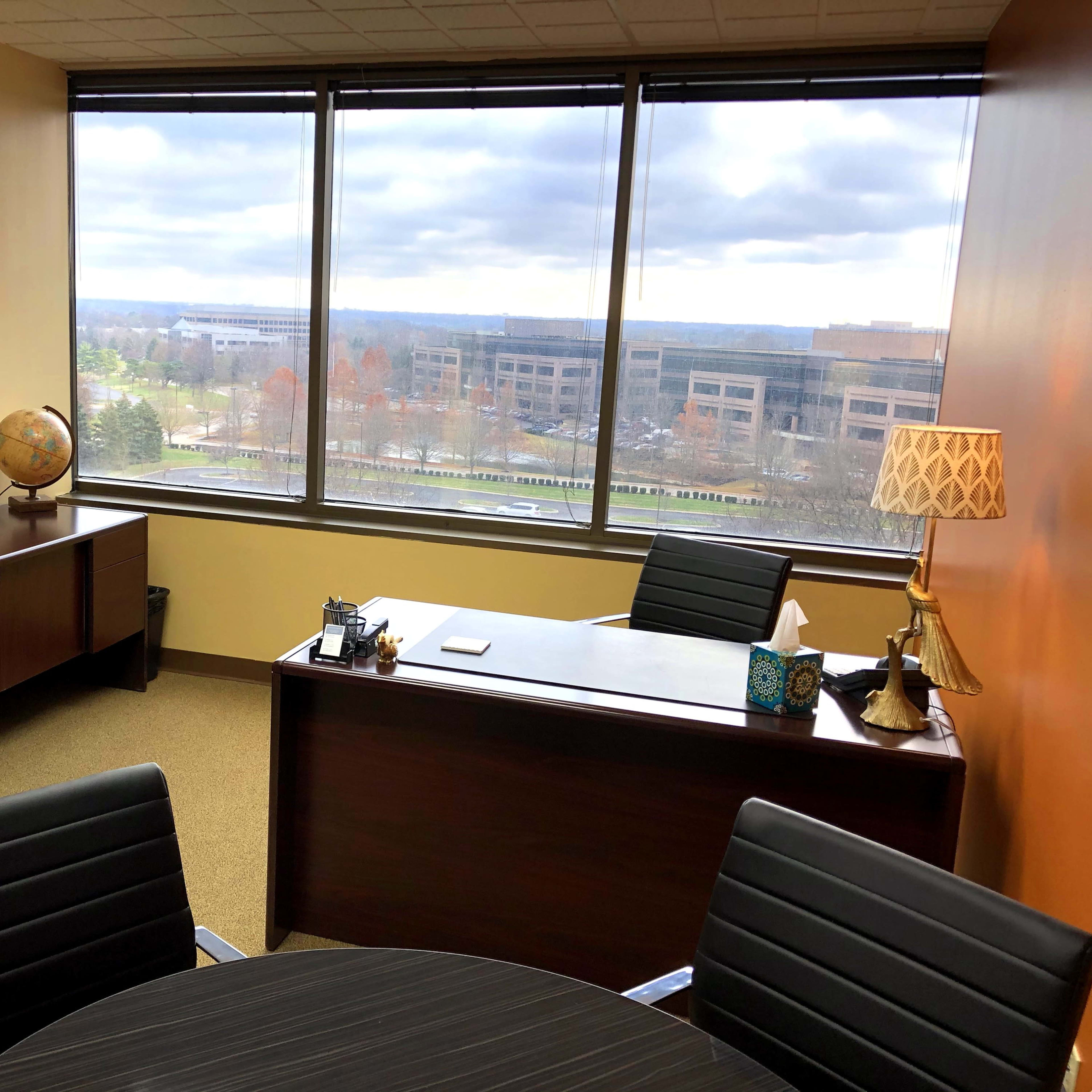 A modern office with a desk, a round table, large windows overlooking a landscape, and decorative items like a globe and a lamp.