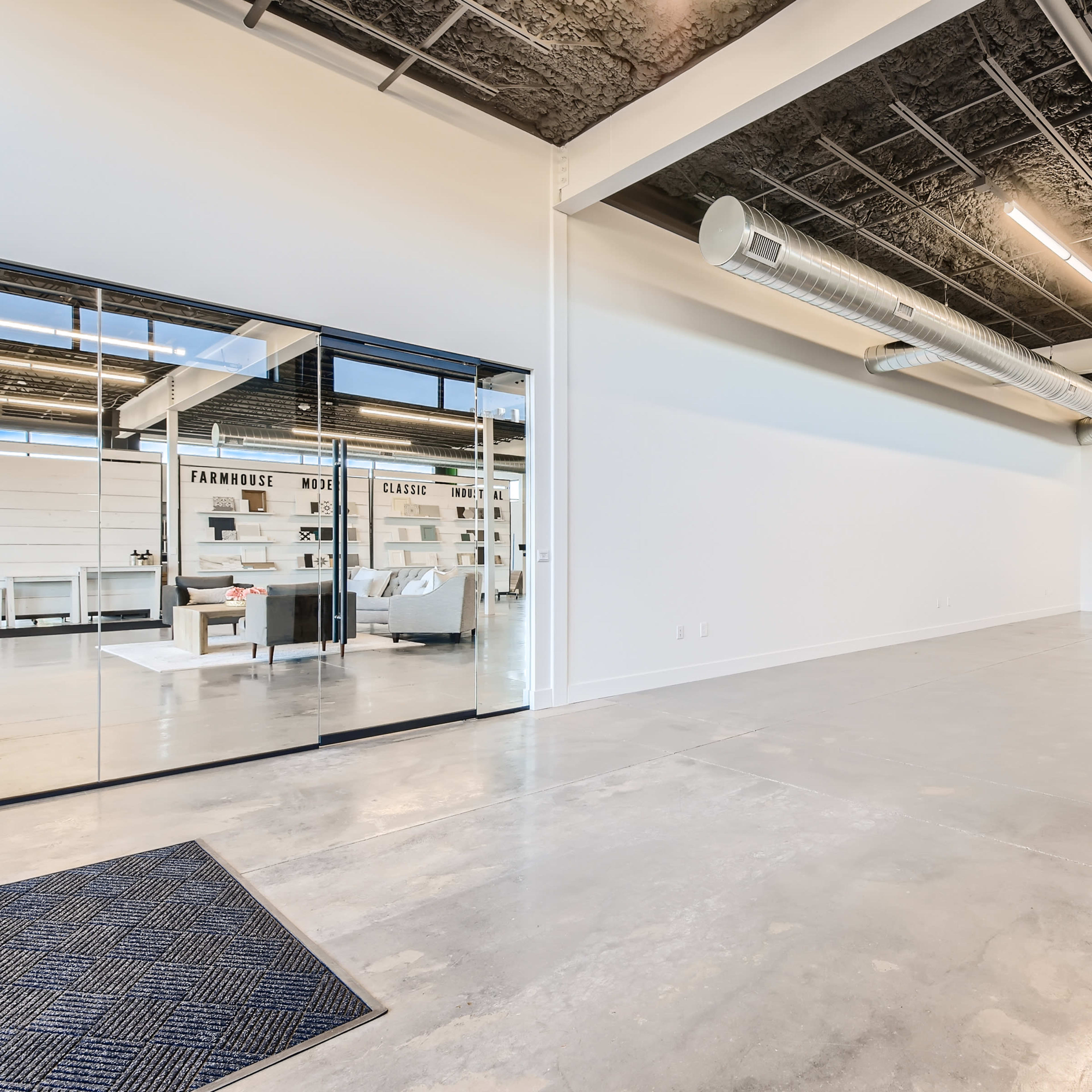 A spacious, empty interior with polished concrete floors and exposed ductwork, leading to a glass-enclosed area featuring furniture and décor.