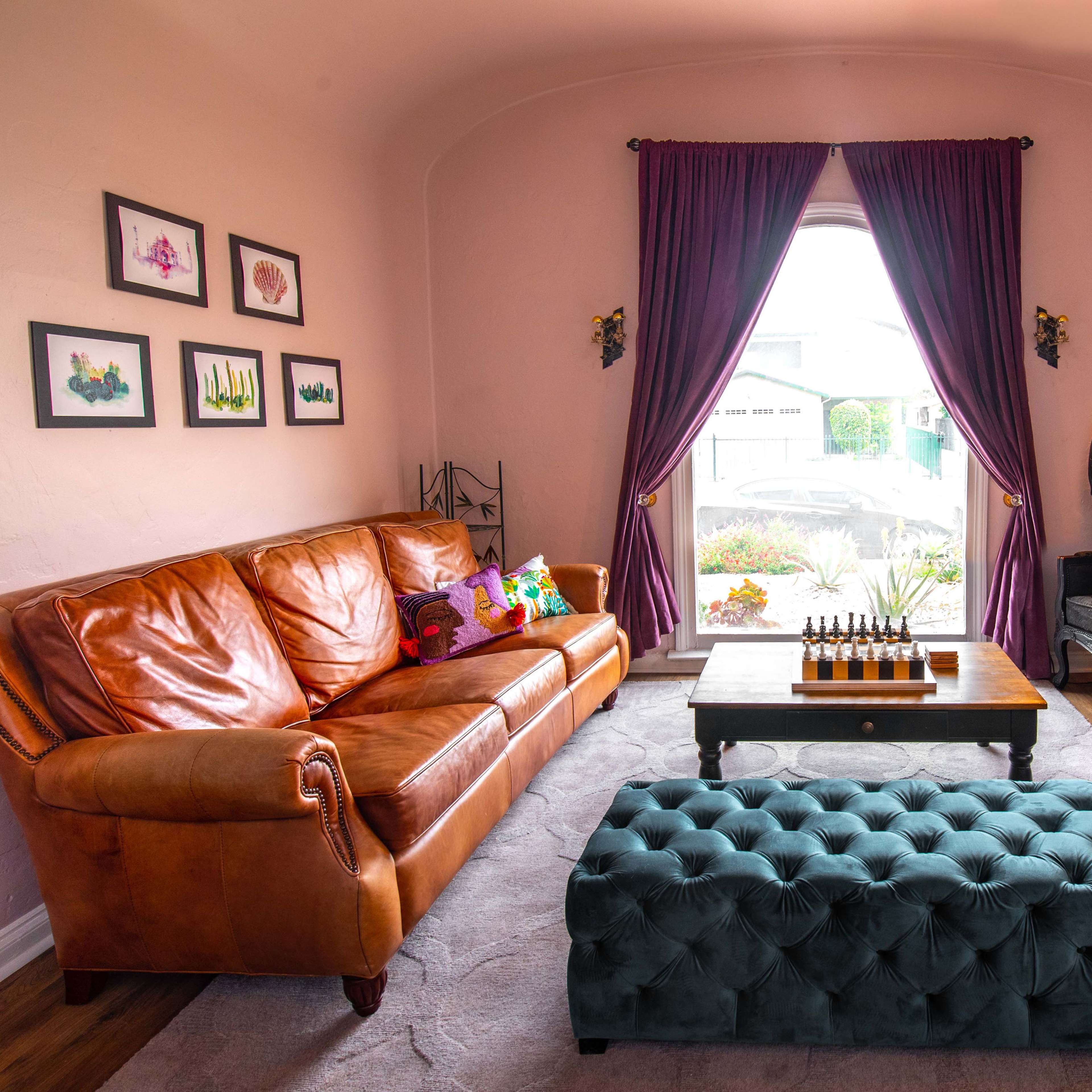 A living room features a brown leather sofa, a tufted teal ottoman, and two large armchairs near a window with purple curtains.
