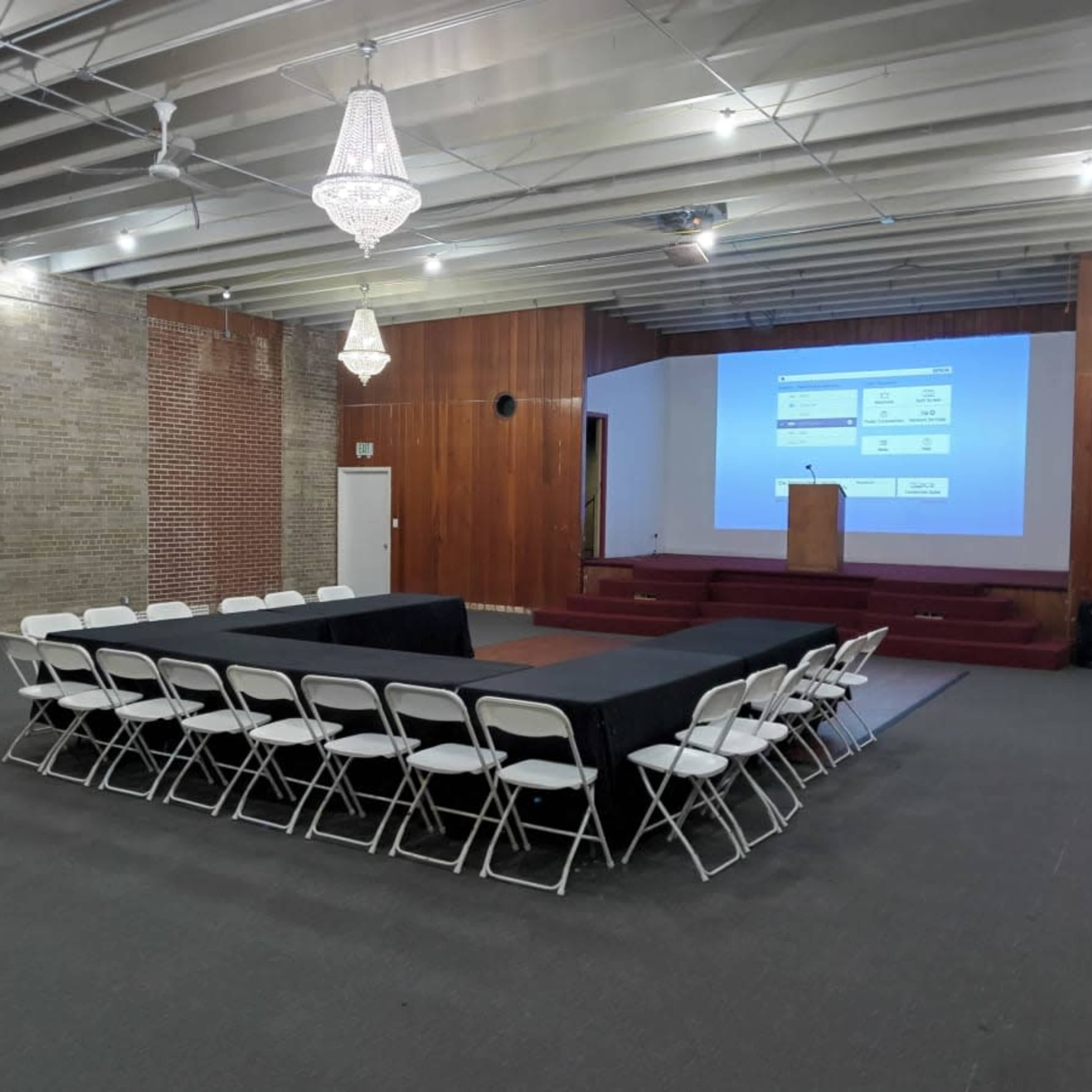 A rectangular arrangement of tables with chairs surrounds a small stage featuring a podium and a large screen in a spacious, well-lit room.