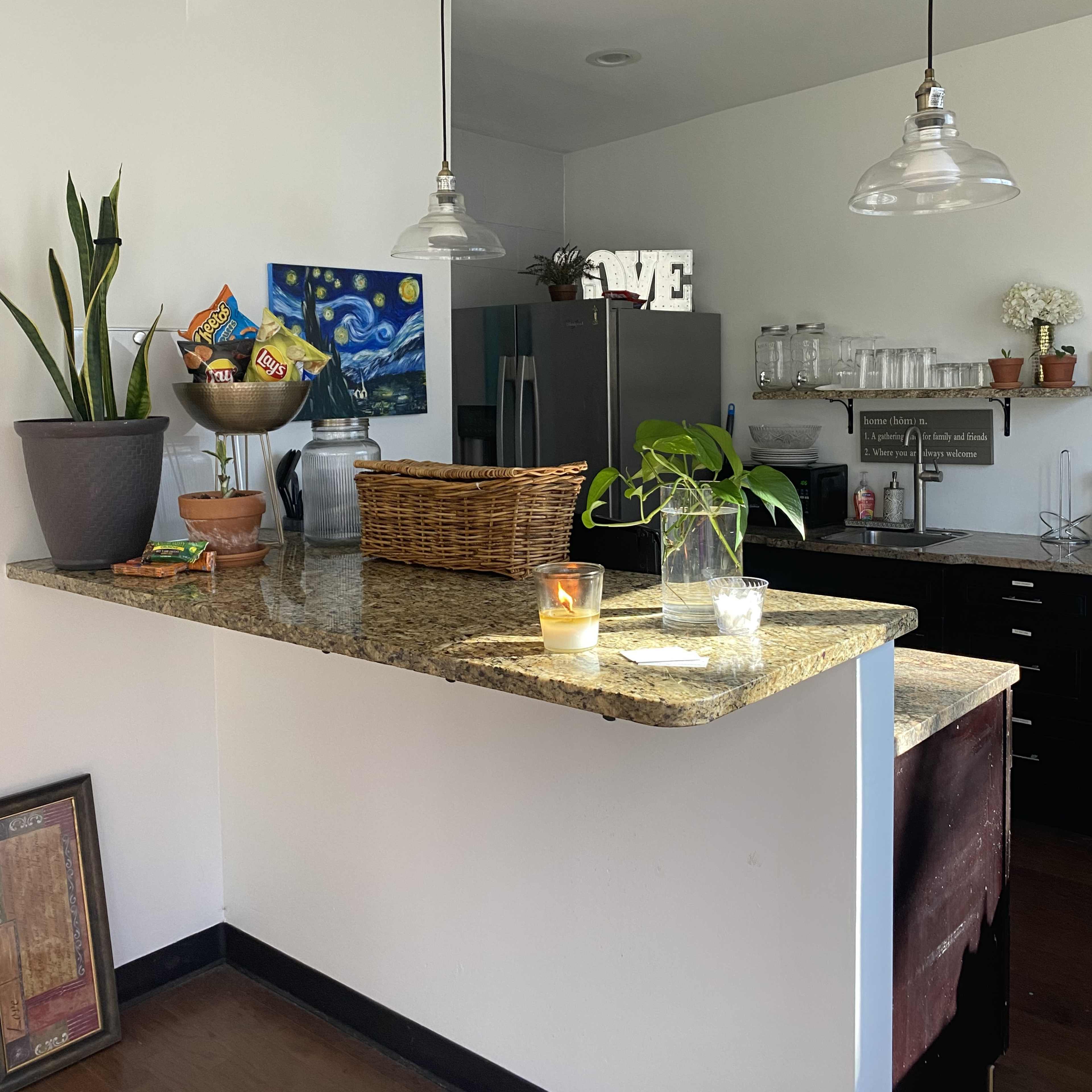 A granite kitchen island is adorned with a basket of snacks, a potted plant, and a candle, while overhead lights illuminate the space.