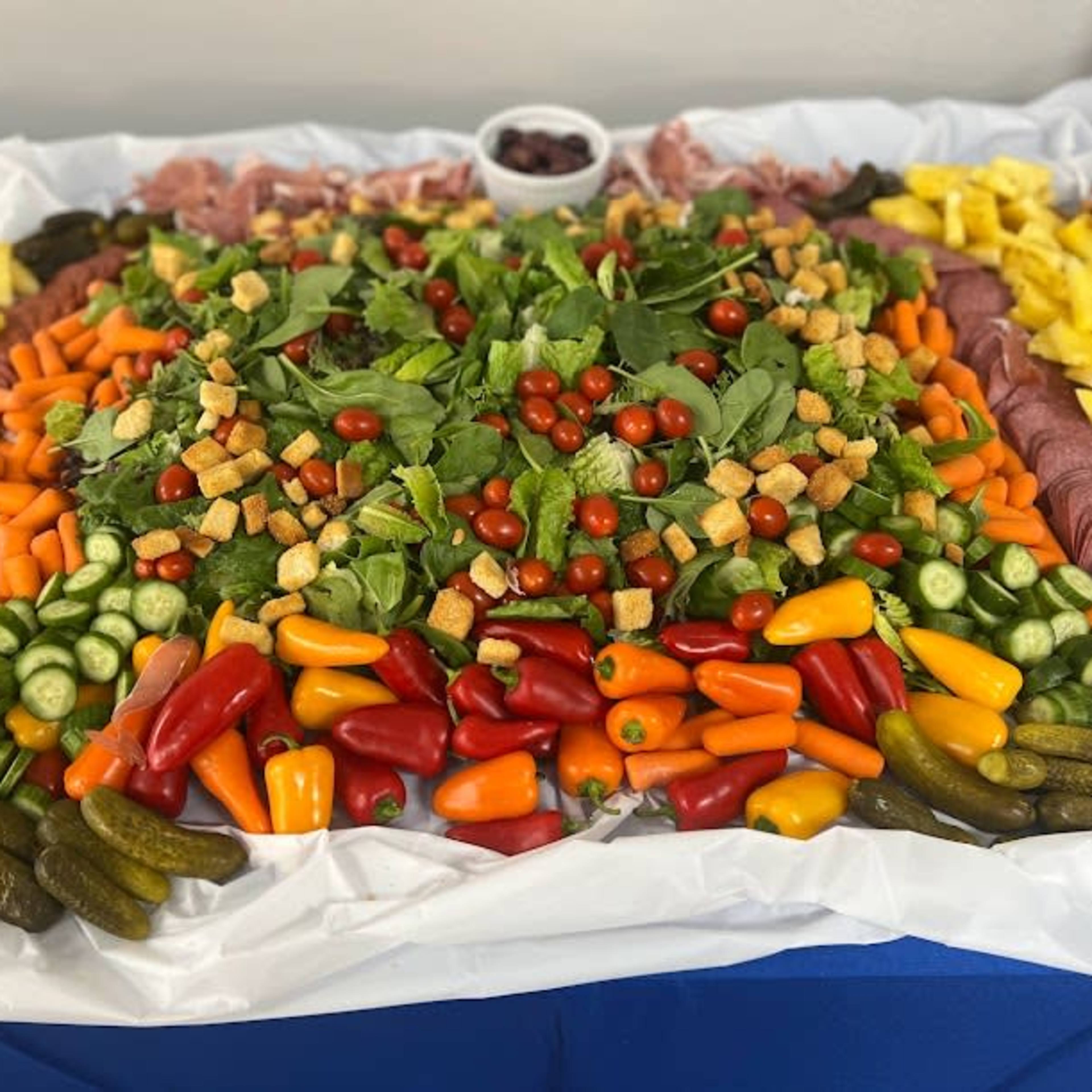 A large platter is arranged with an assortment of salad greens, cherry tomatoes, assorted peppers, pickles, olives, and various fruits, surrounded by bowls of grapes and crackers.