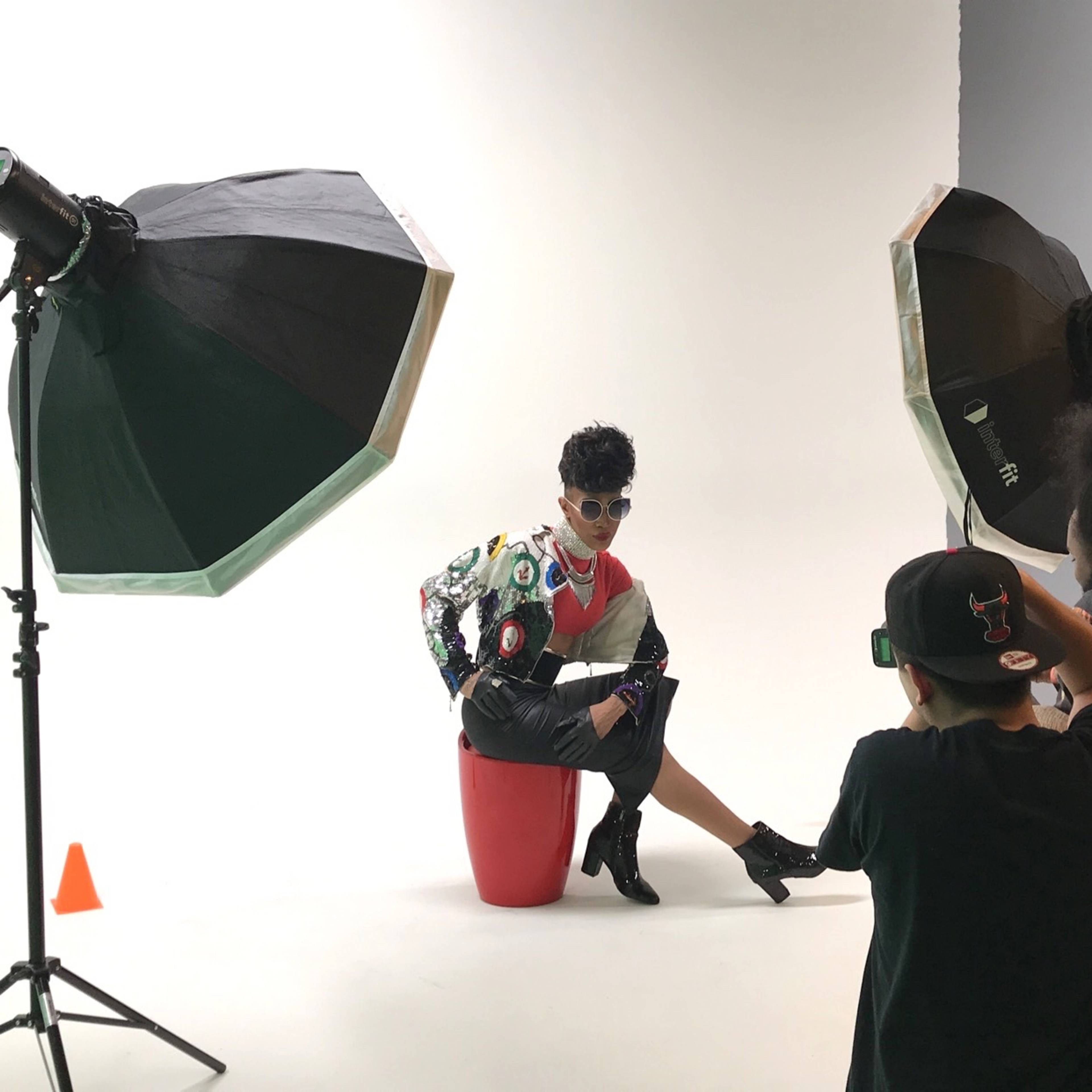A model poses on a red stool in front of a white backdrop while photographers adjust lighting equipment.