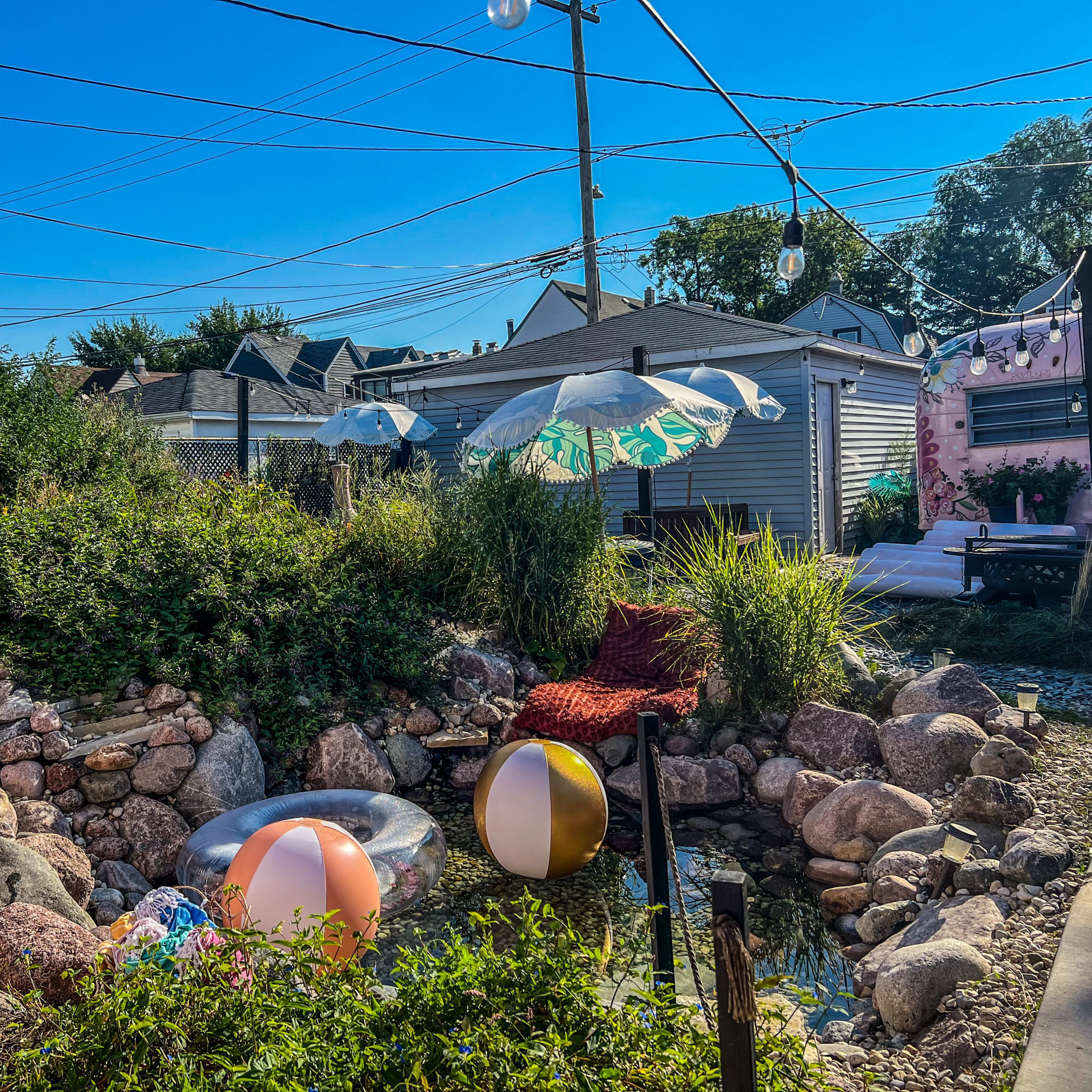 A backyard scene features a rock garden with beach balls, umbrellas, and string lights, set against a clear blue sky.