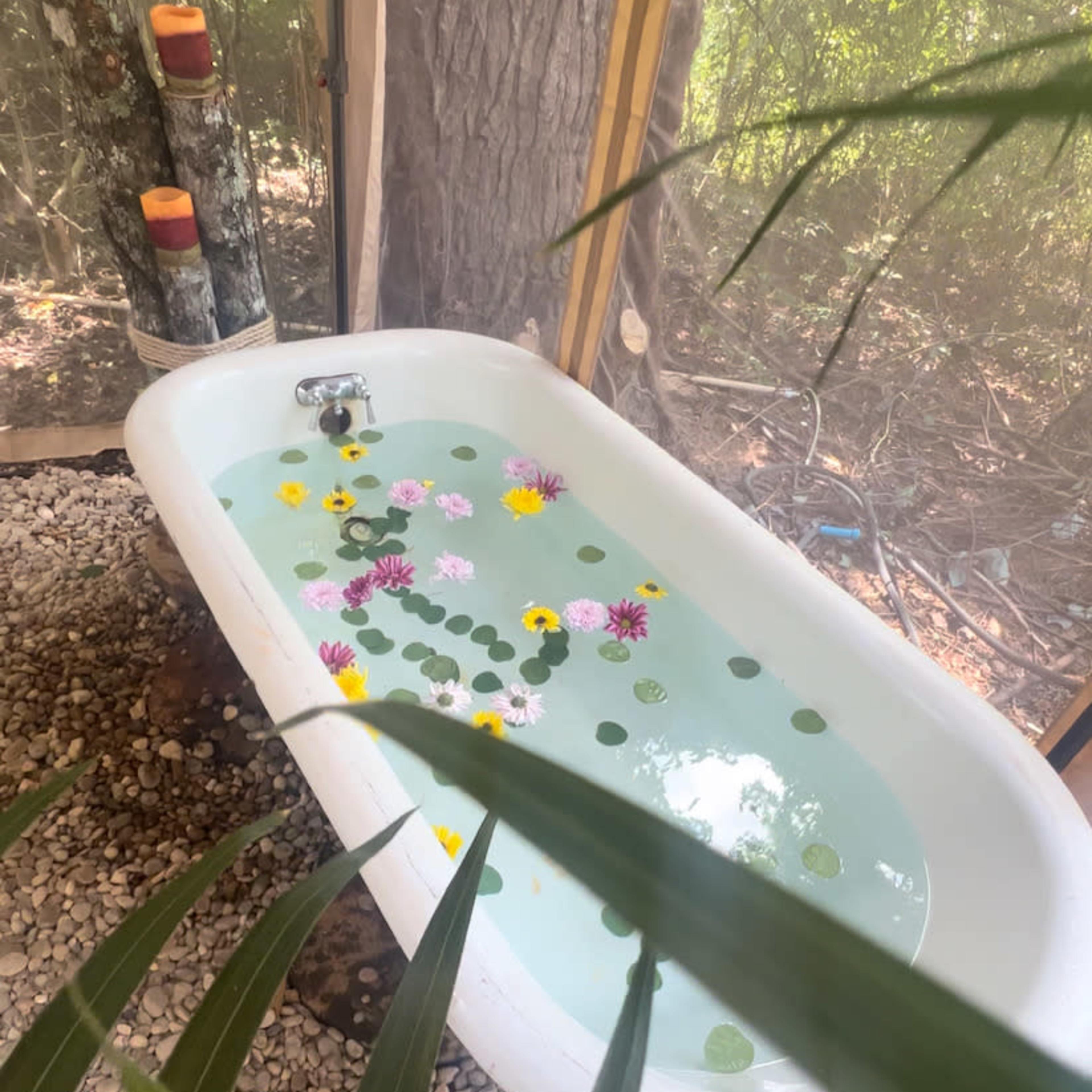 A freestanding bathtub filled with water and floating flowers is positioned among pebbles and surrounded by greenery.