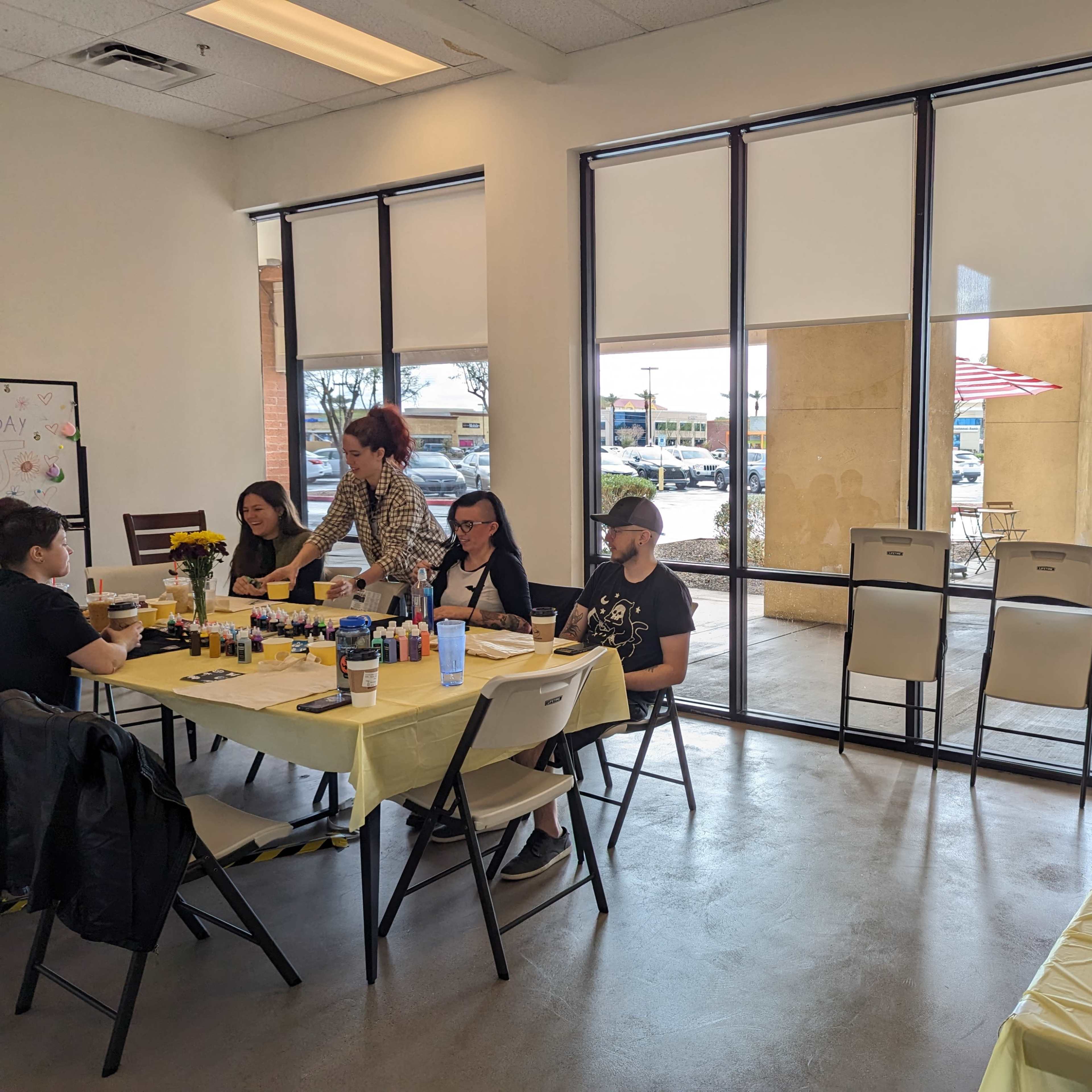 A brightly lit room with a table set up for a crafting event, featuring people engaged in activities and a display of decorated cupcakes on one side.