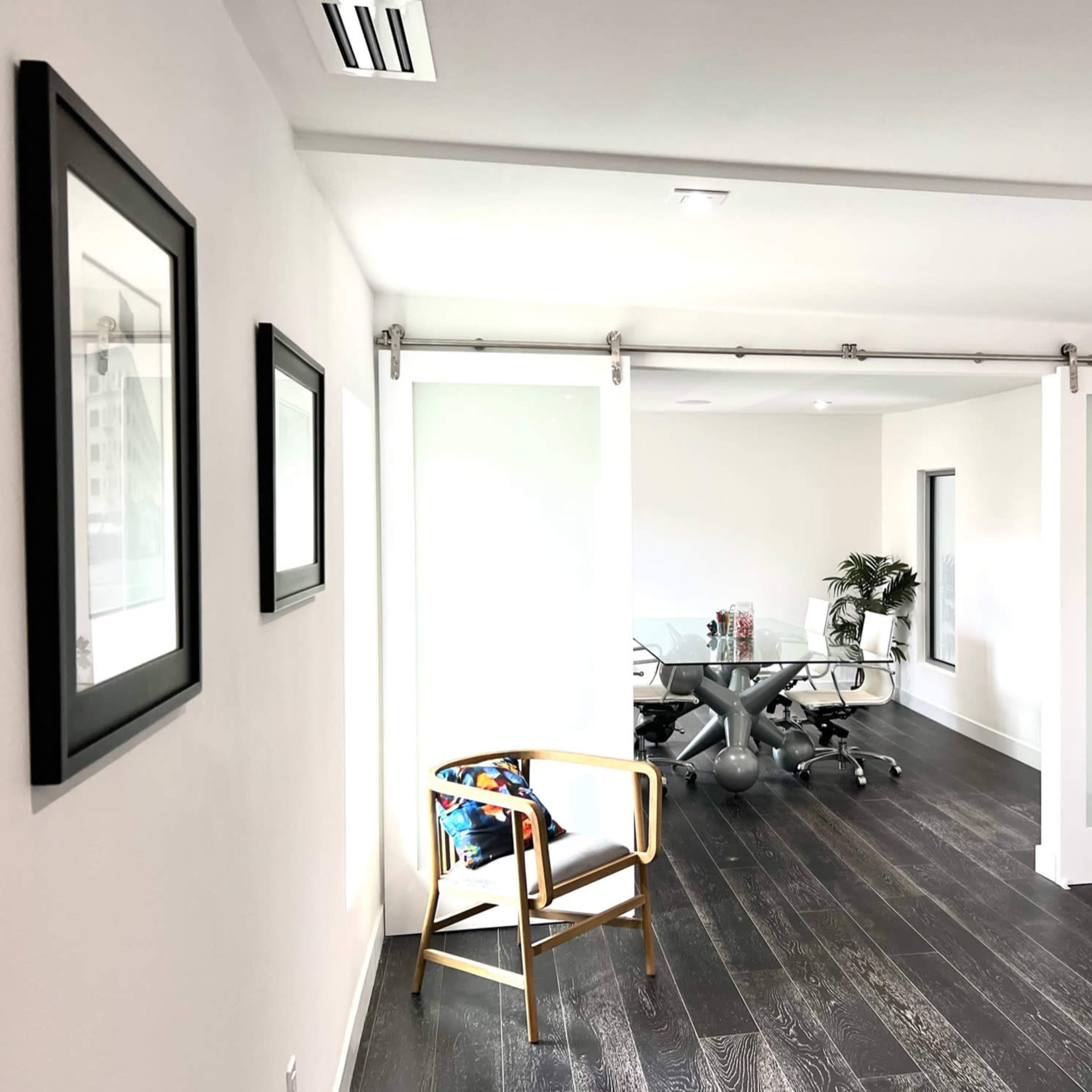 A modern office space featuring a glass conference table and a sliding door, with framed artwork on the walls and dark wood flooring.