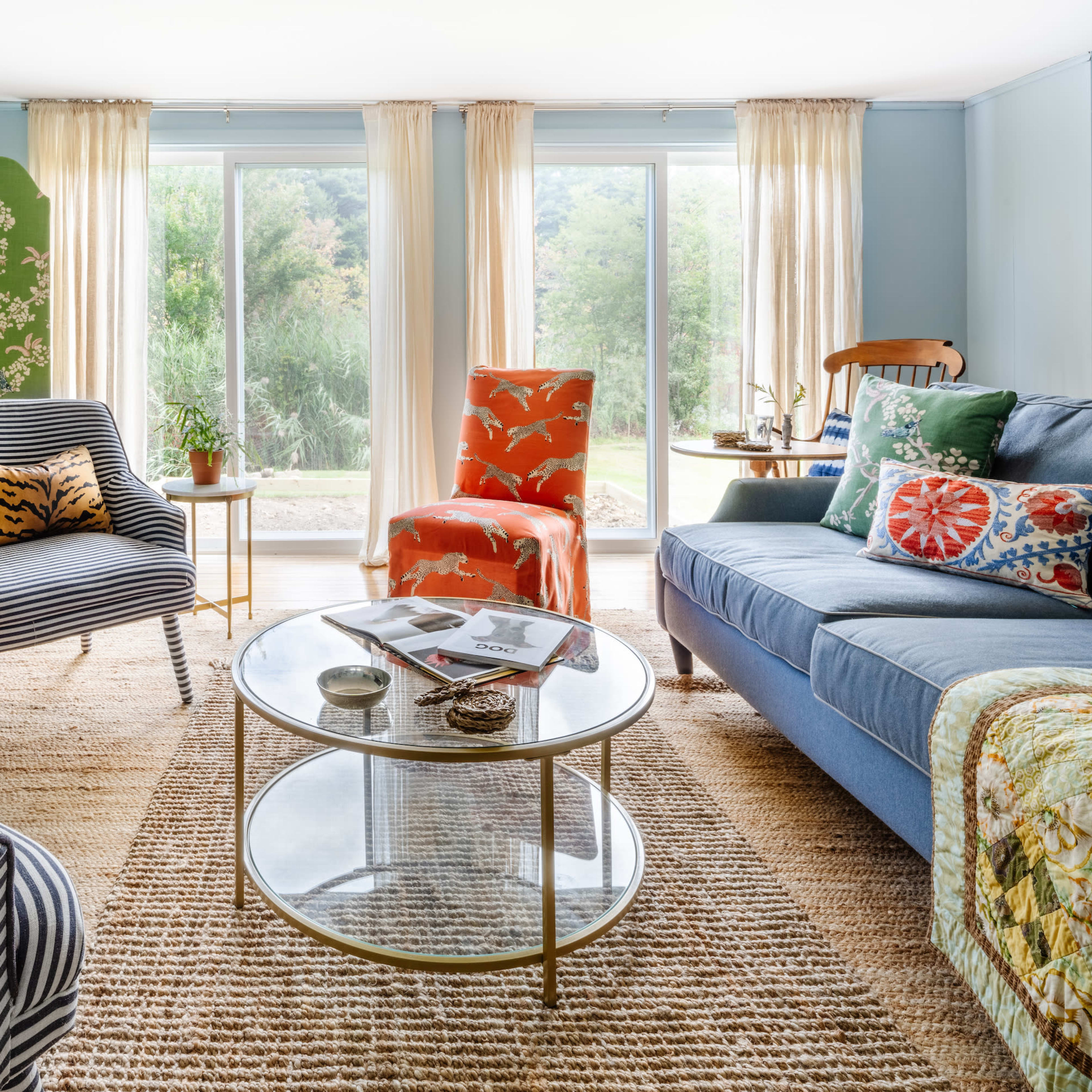 A living room features a blue sofa, patterned armchairs, a round coffee table, and large windows overlooking a garden.