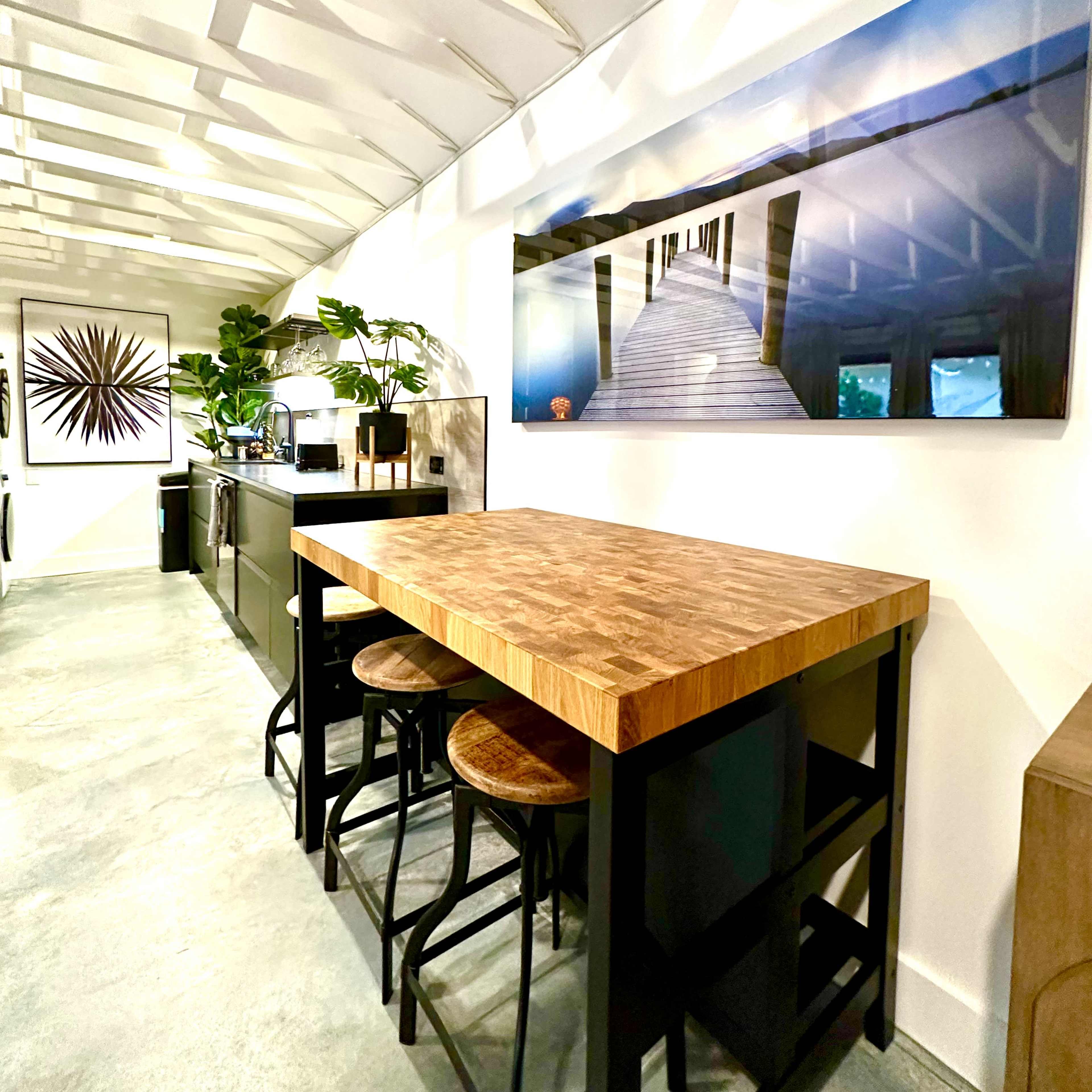 A modern kitchen with a wooden table and stools, framed by white walls and a textured ceiling with plants and artwork on display.