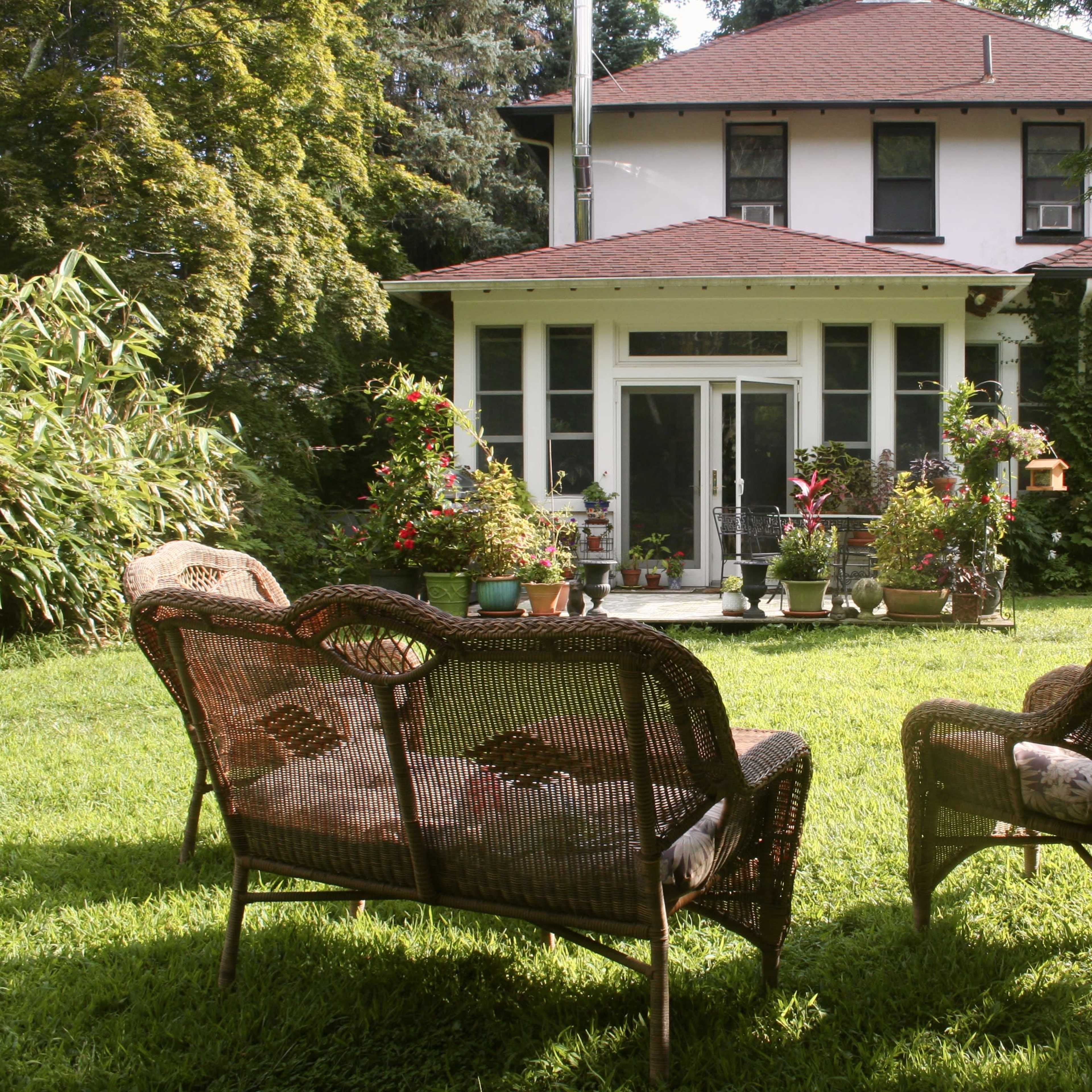 A garden with two wicker chairs in the foreground, facing a white house with a red roof and surrounded by greenery.
