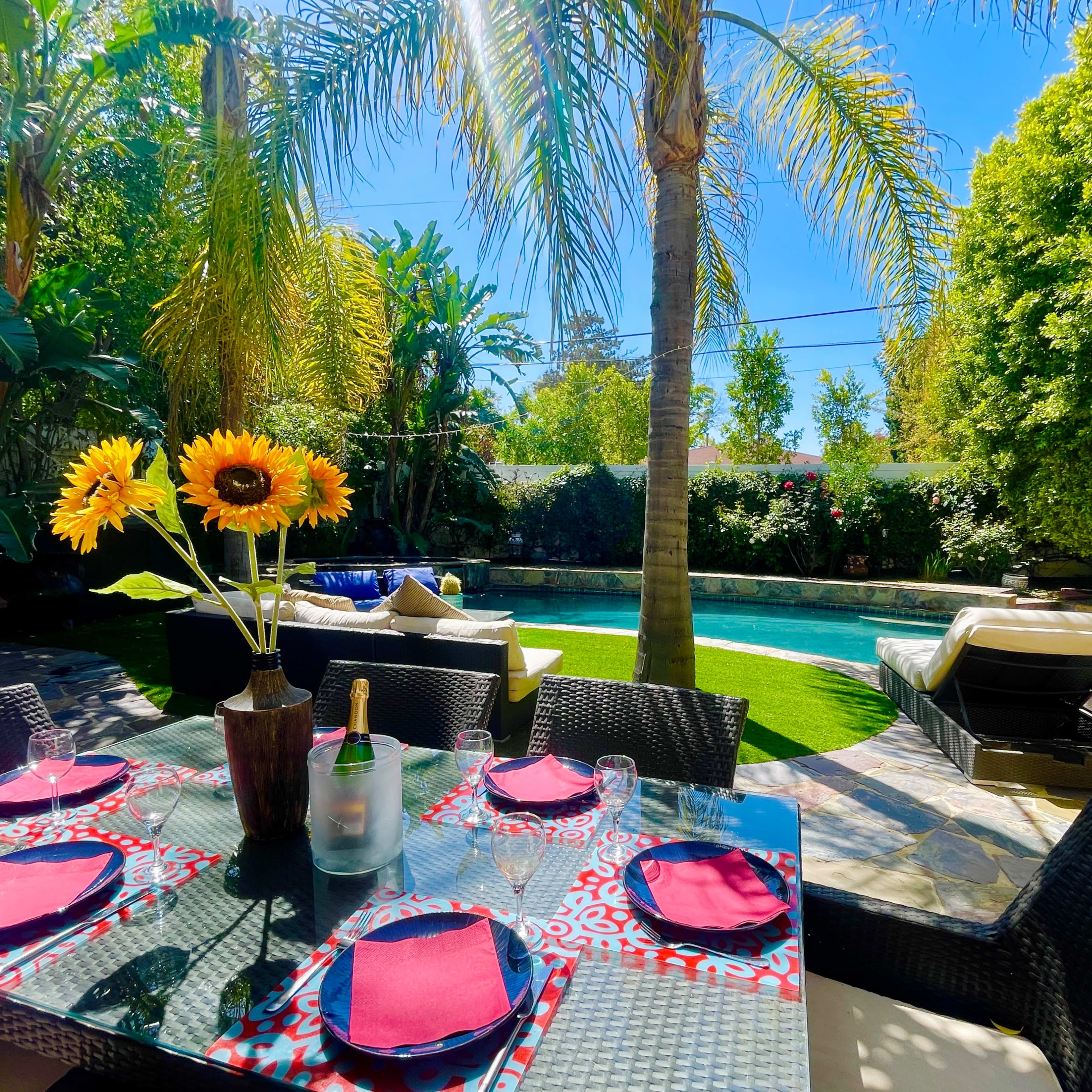 A dining table with sunflowers and champagne overlooks a swimming pool surrounded by palm trees and lush greenery.