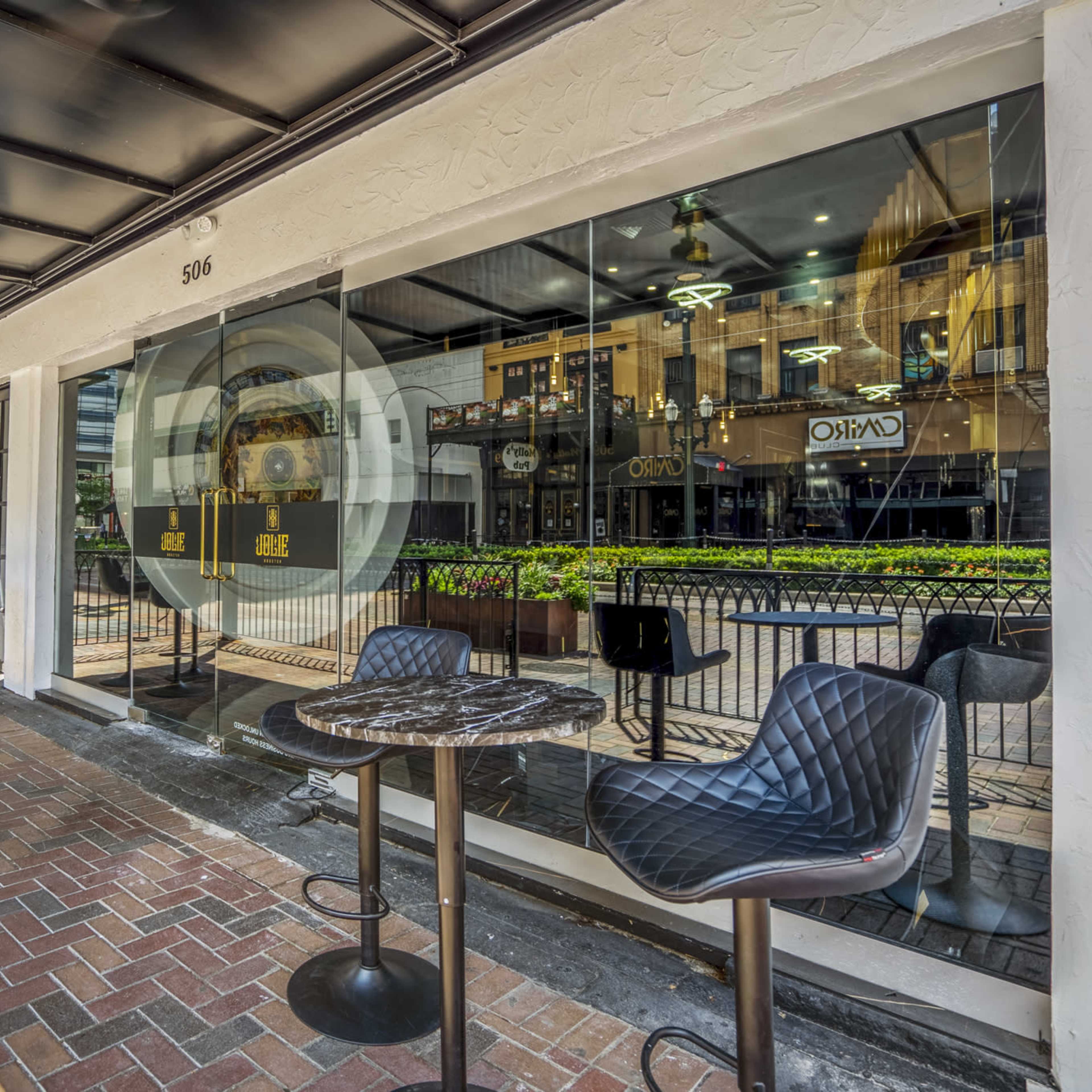 An outdoor dining area with black chairs and small tables in front of a glass storefront, surrounded by greenery and a walkway.