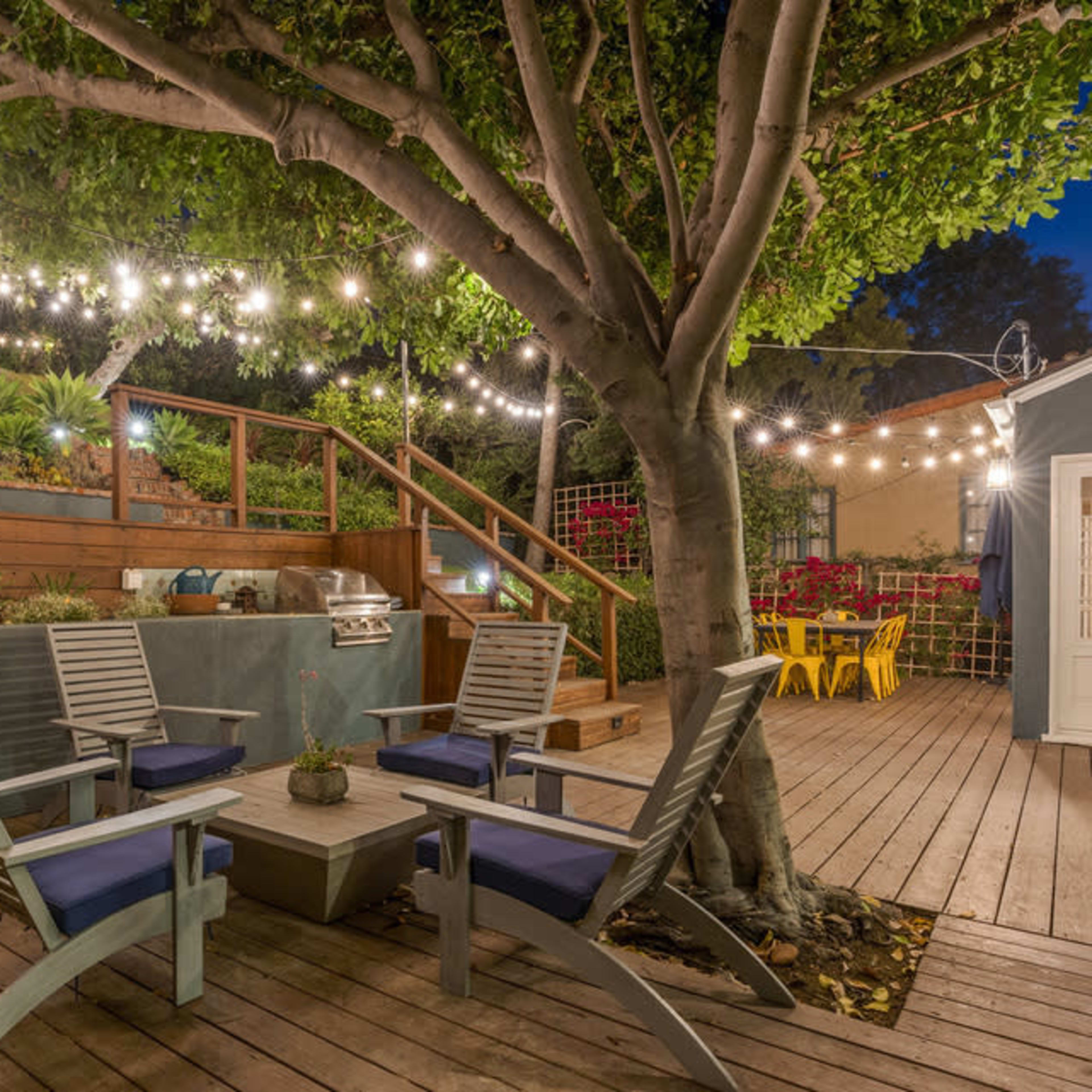 A cozy outdoor patio area features wooden chairs arranged around a table under string lights, with a staircase leading to a landscaped garden.