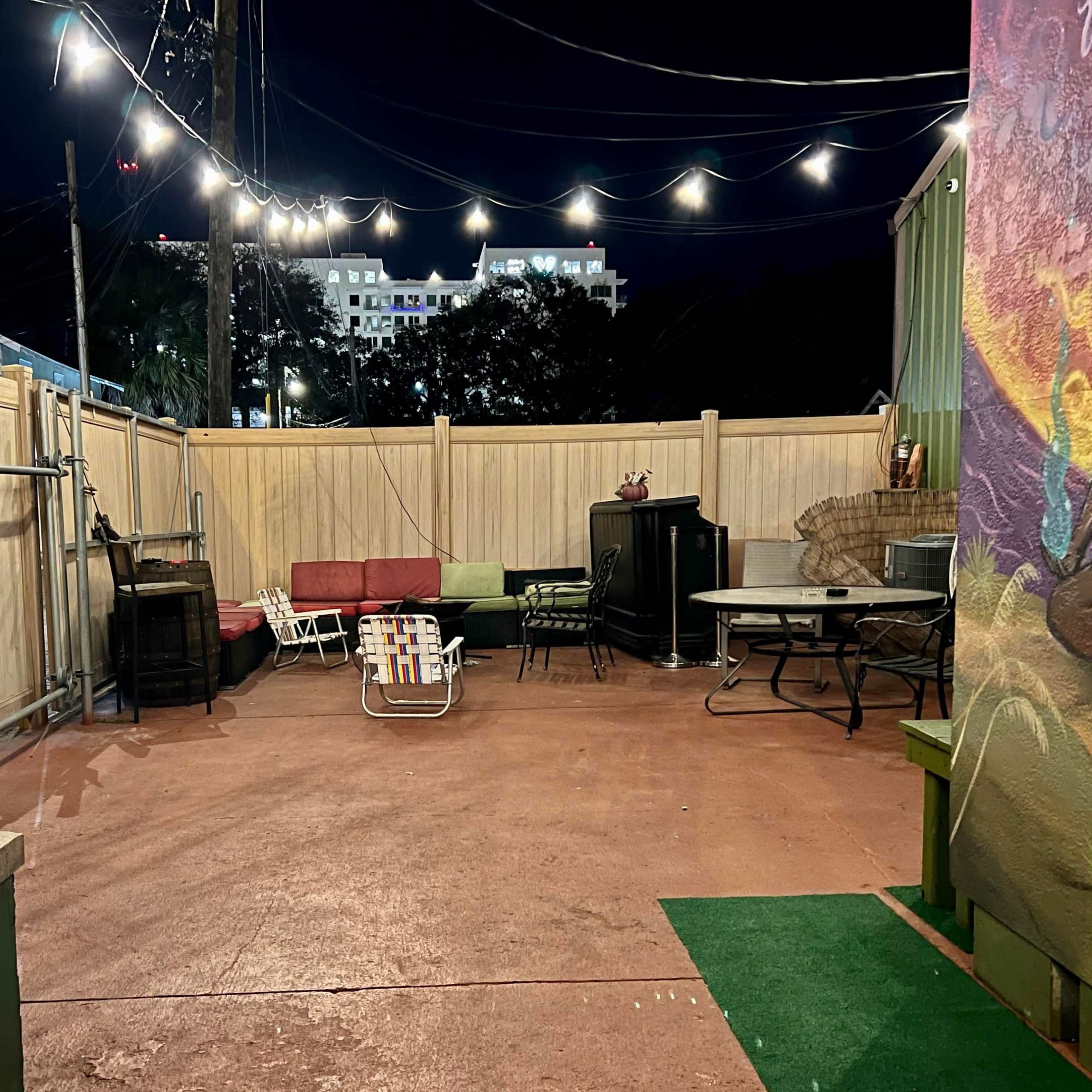 An outdoor patio area at night, featuring a seating arrangement with a red sofa, a folding chair, a dining set, and a wall mural.