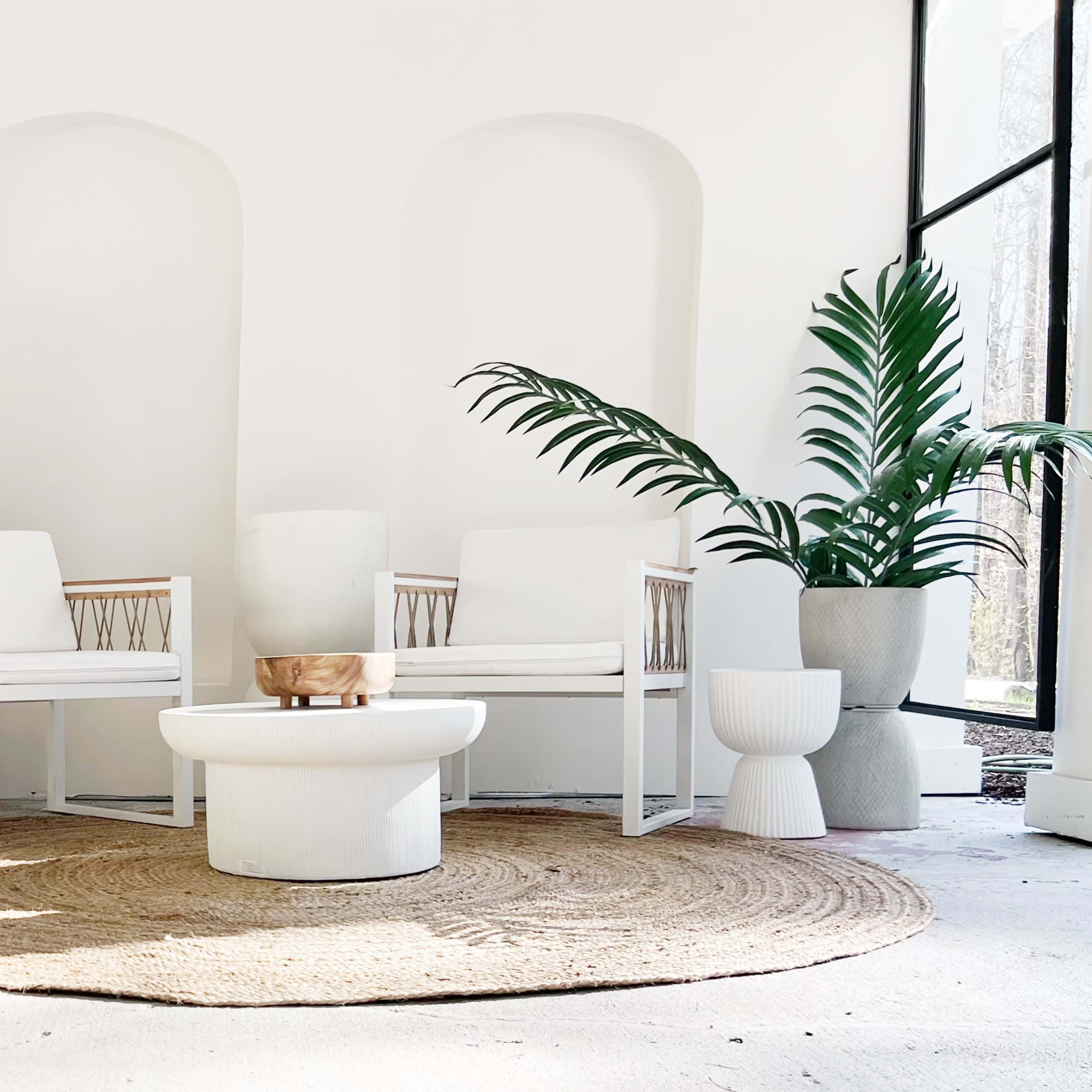 A seating area features white chairs and tables arranged around a circular woven rug, with decorative plant elements and textured vases against a light backdrop.