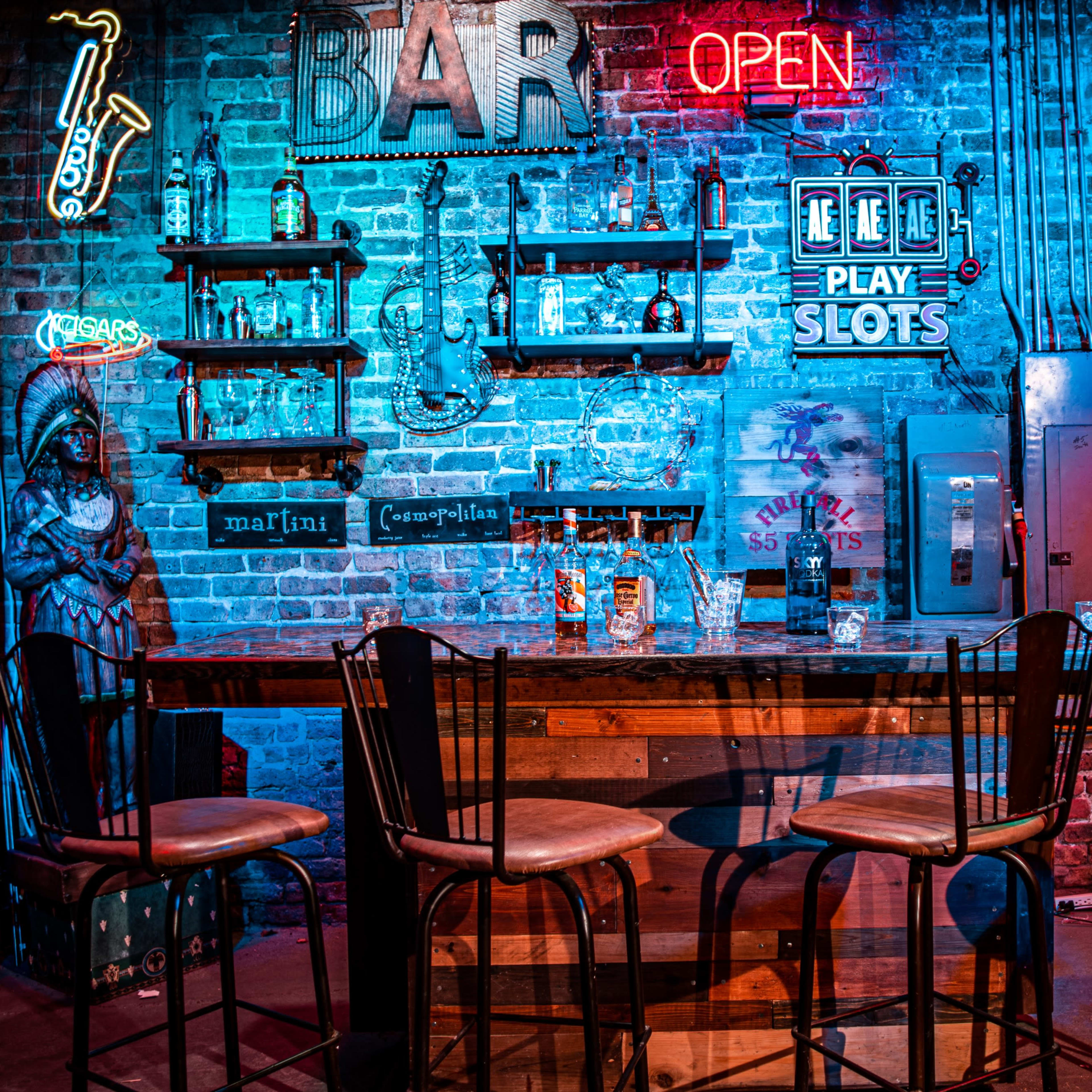 A dimly lit bar scene features a brick wall adorned with neon signs, shelves of liquor bottles, and wooden bar stools arranged around a large wooden bar.