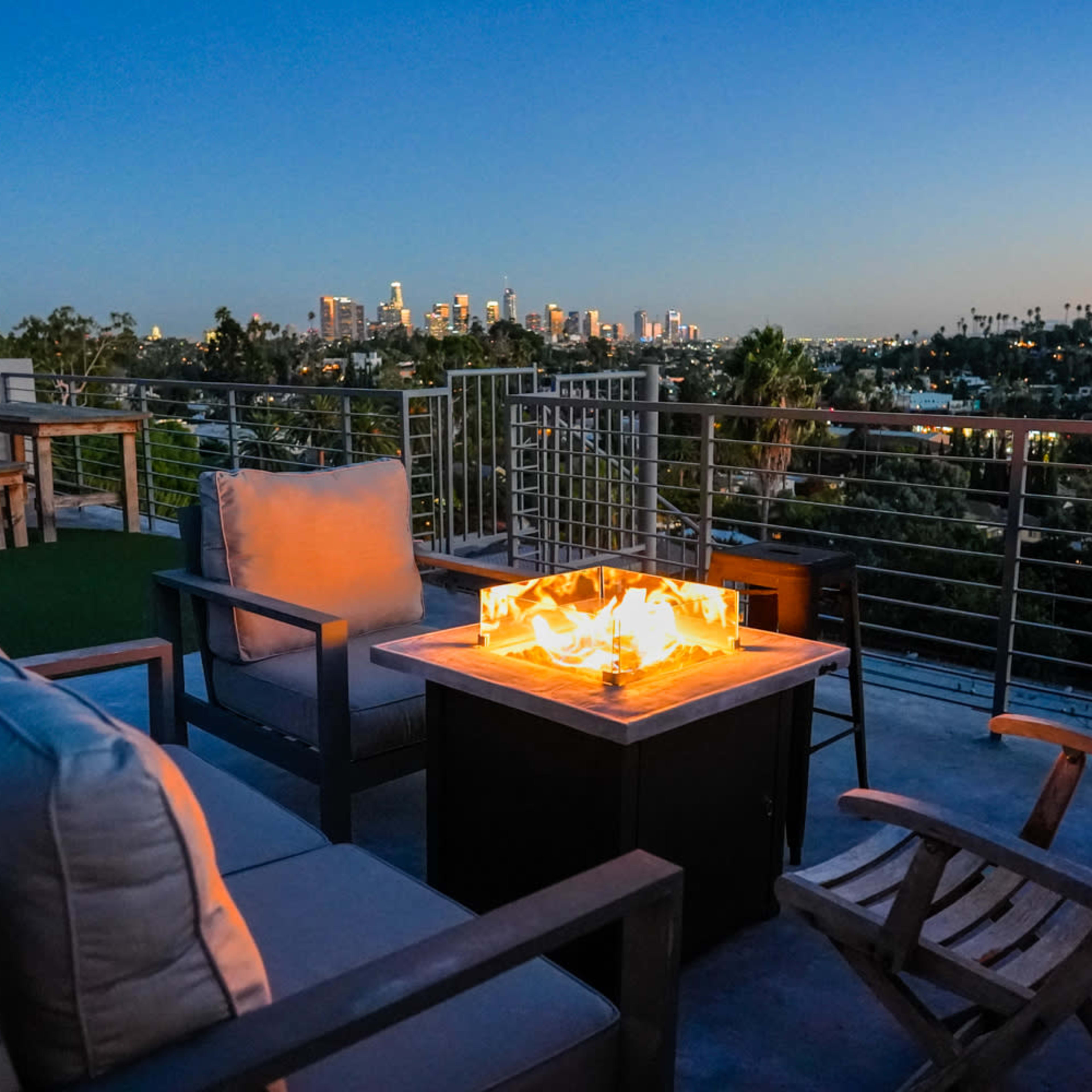 A rooftop lounge area features comfortable seating and a fire pit, with a city skyline visible in the background at dusk.