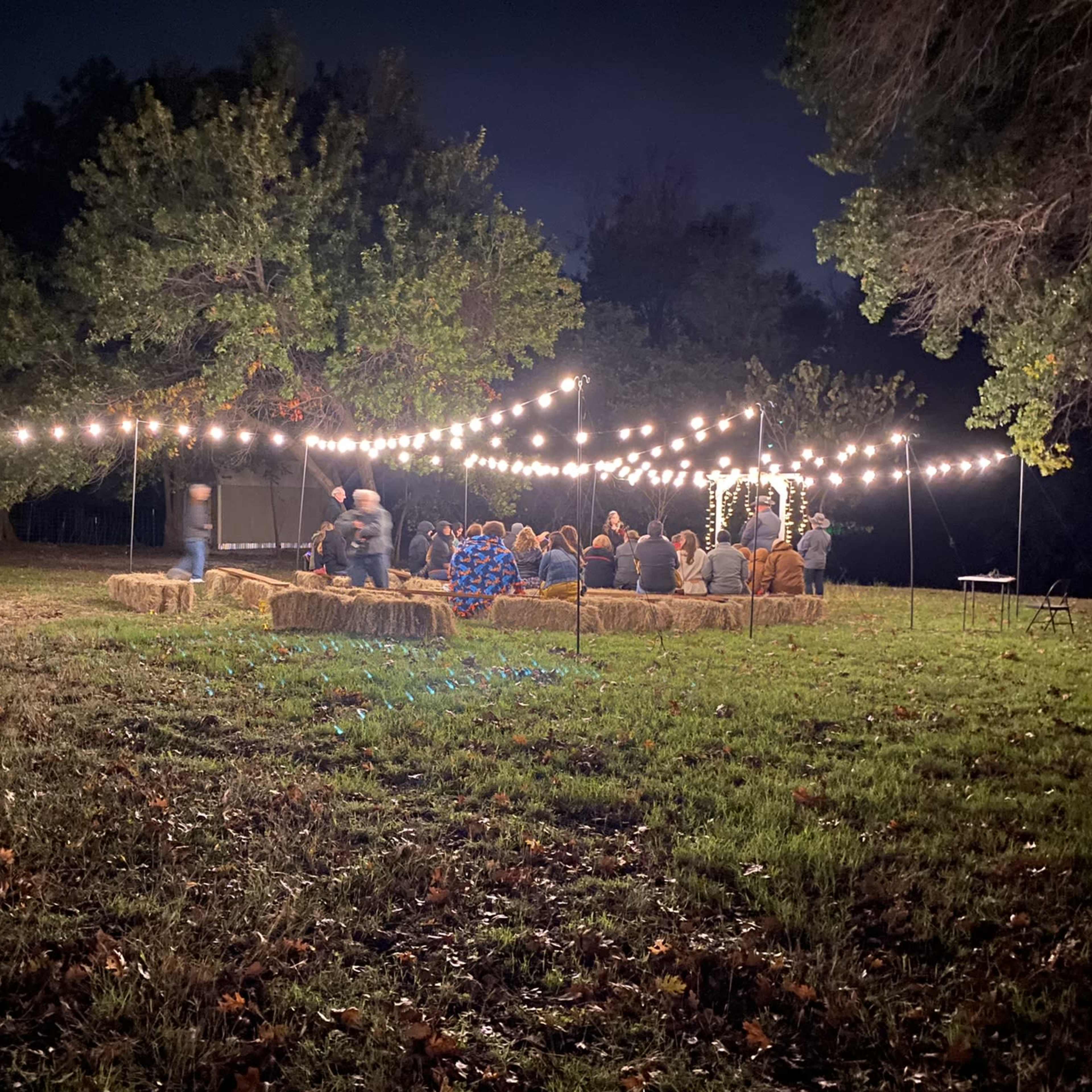 A group of people sits in a grassy area beneath strings of lights, while candles are arranged on a barrel in the foreground.