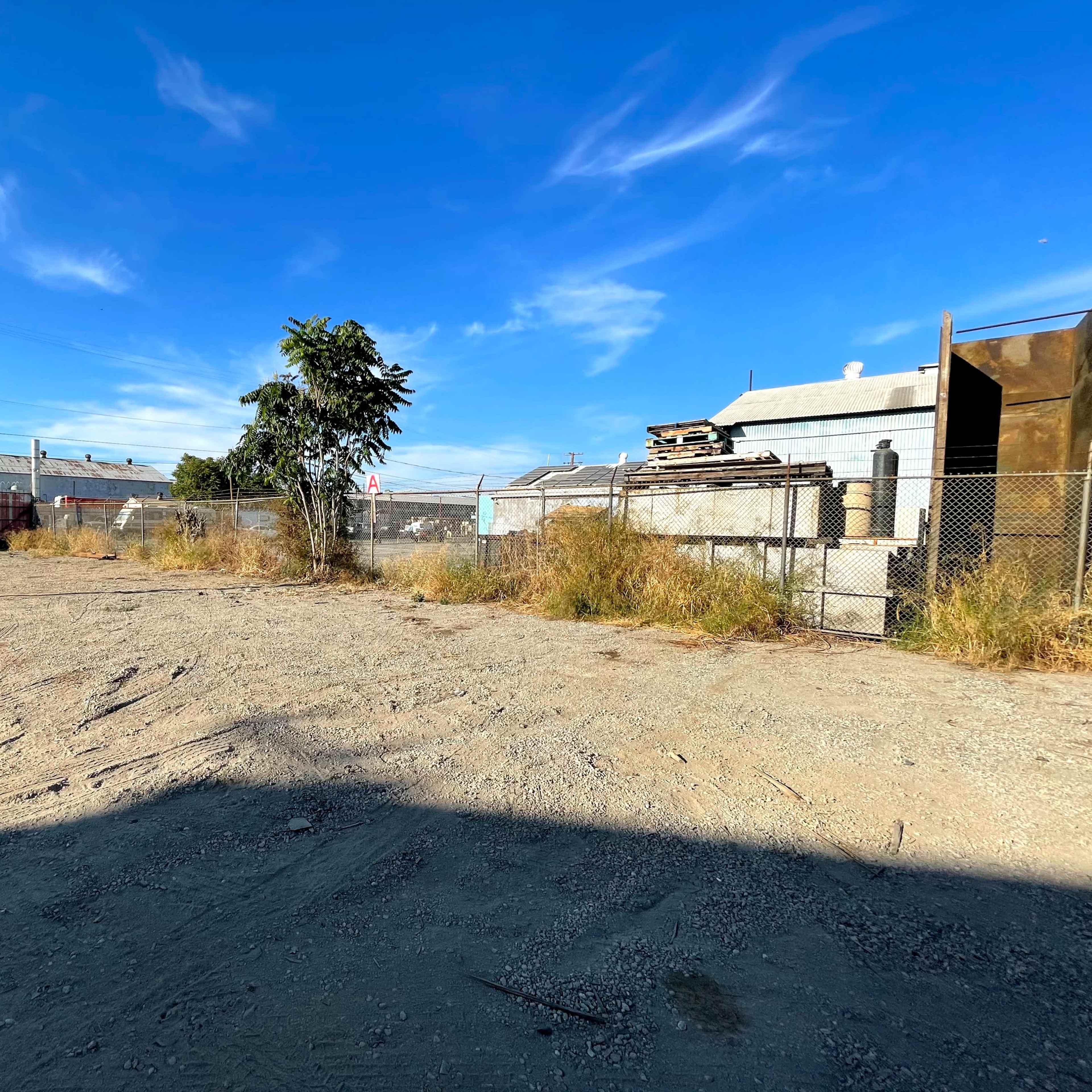 A dusty, vacant lot with overgrown grass and a lone tree, flanked by storage buildings and a chain-link fence.