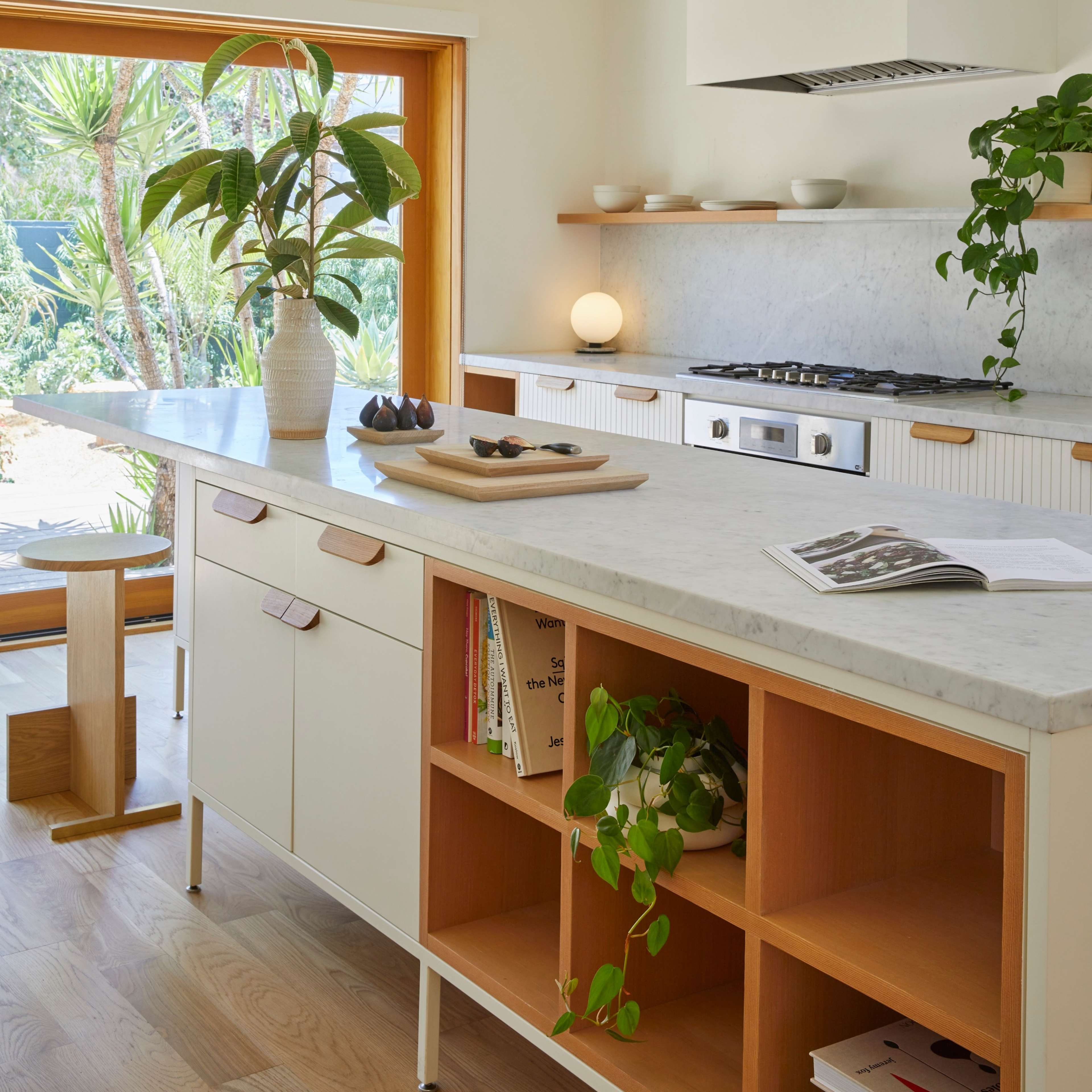 A modern kitchen features a marble countertop, wooden shelves, and plants beside a large window.