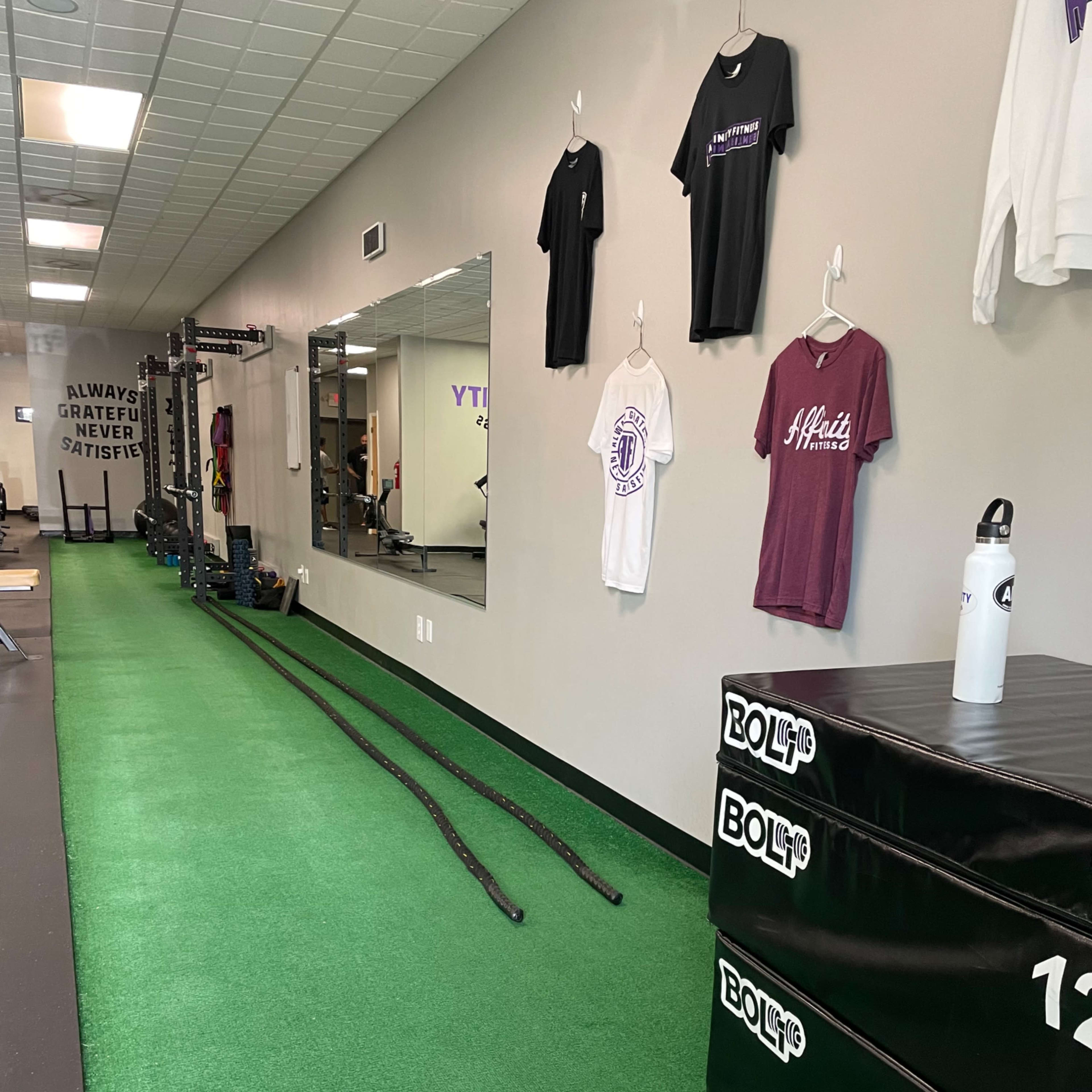 A gym interior with green turf, stacked weight boxes, exercise ropes, and workout gear hanging on the walls.
