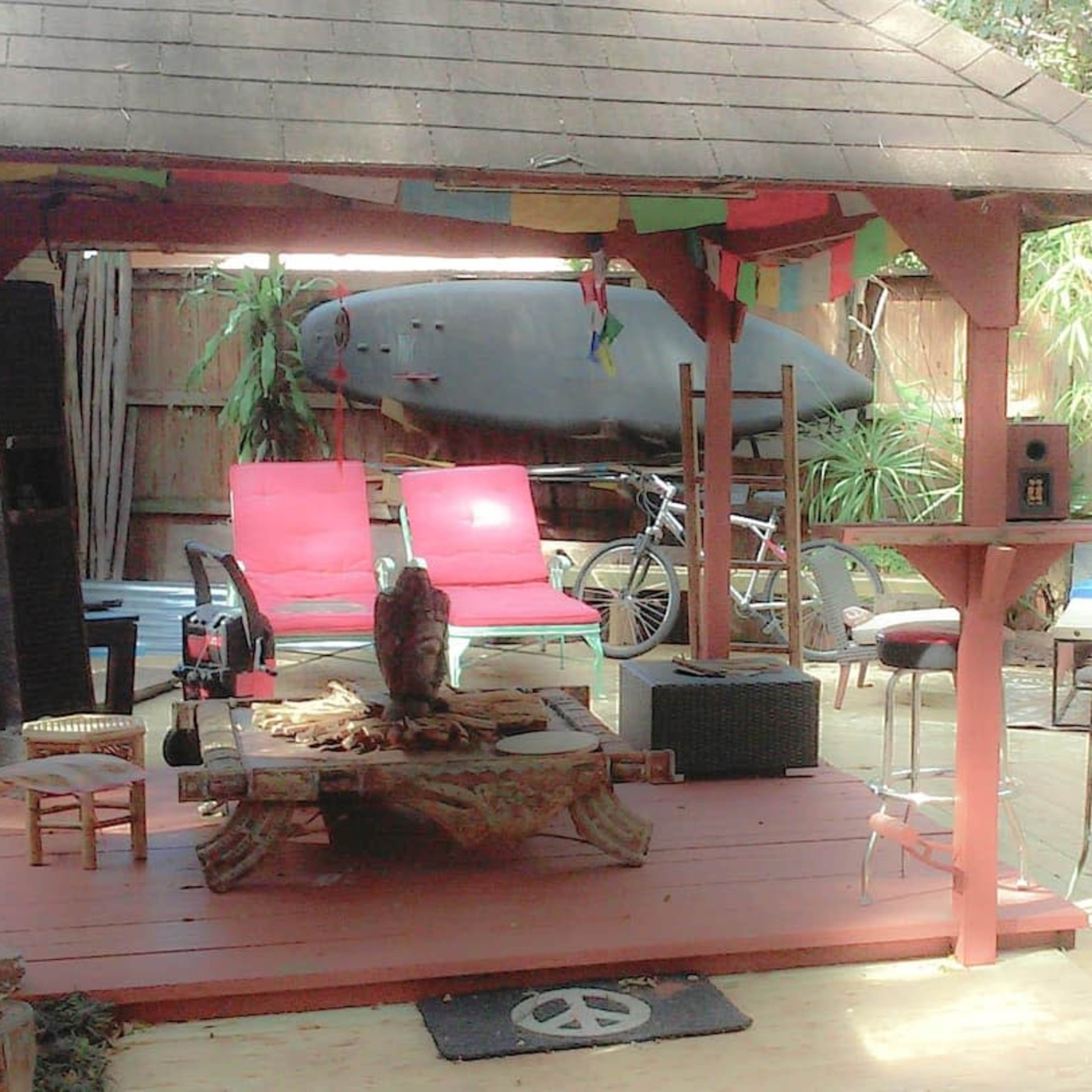 A wooden deck with a shaded seating area featuring red chairs, a low table, and decorative plants, alongside a surfboard mounted in an outdoor shed.